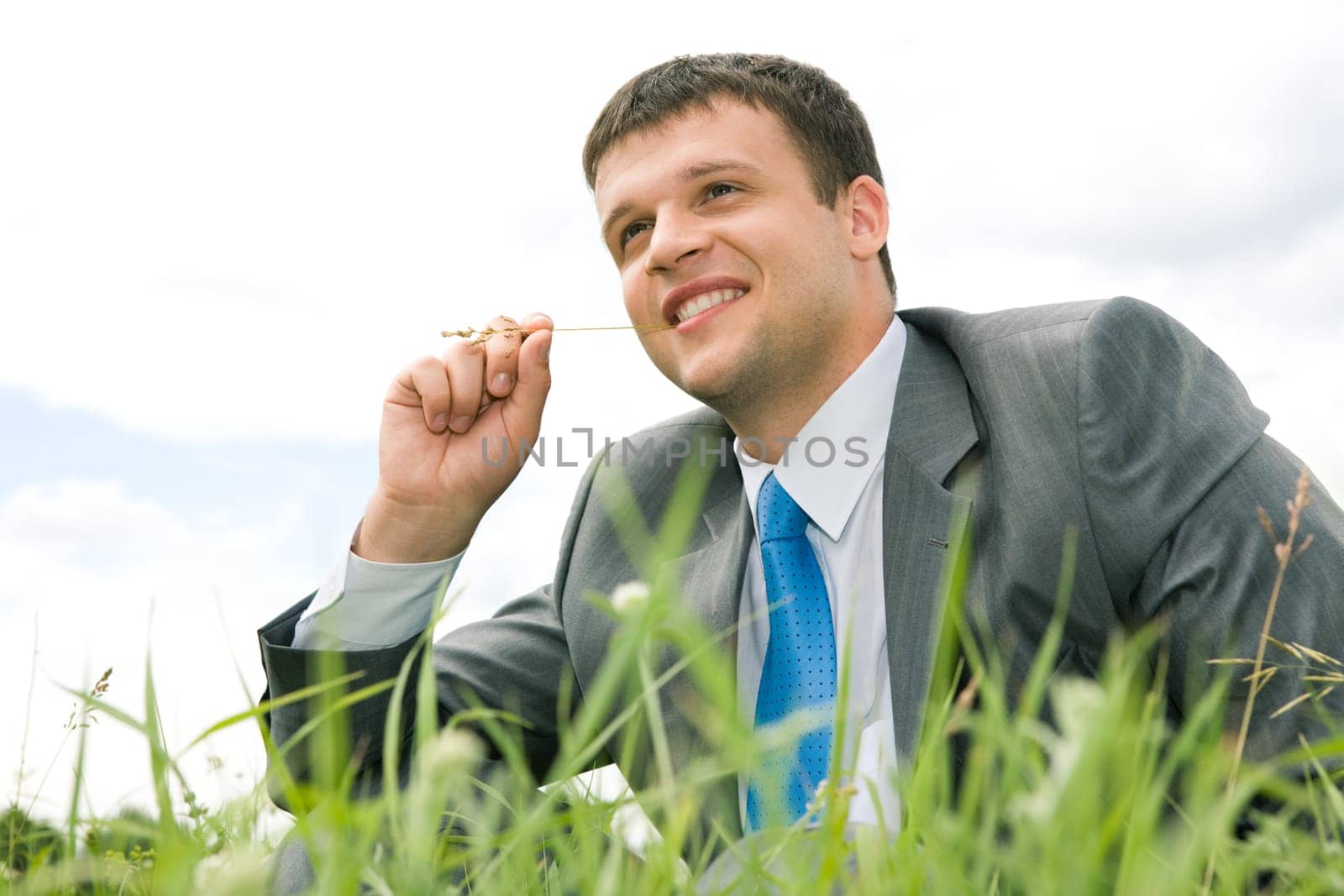 Portrait of cheerful young man sitting on grass with blade in mouth and smiling