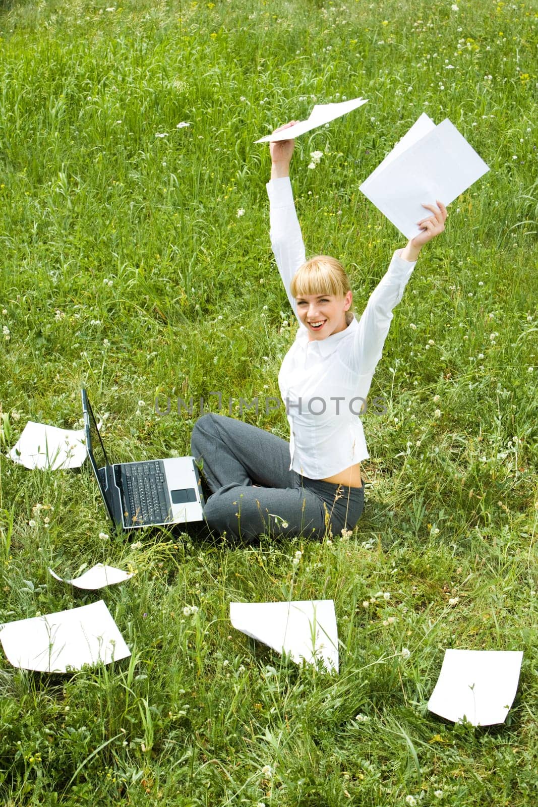 Portrait of joyful female sitting on green grass with raised arms during her work