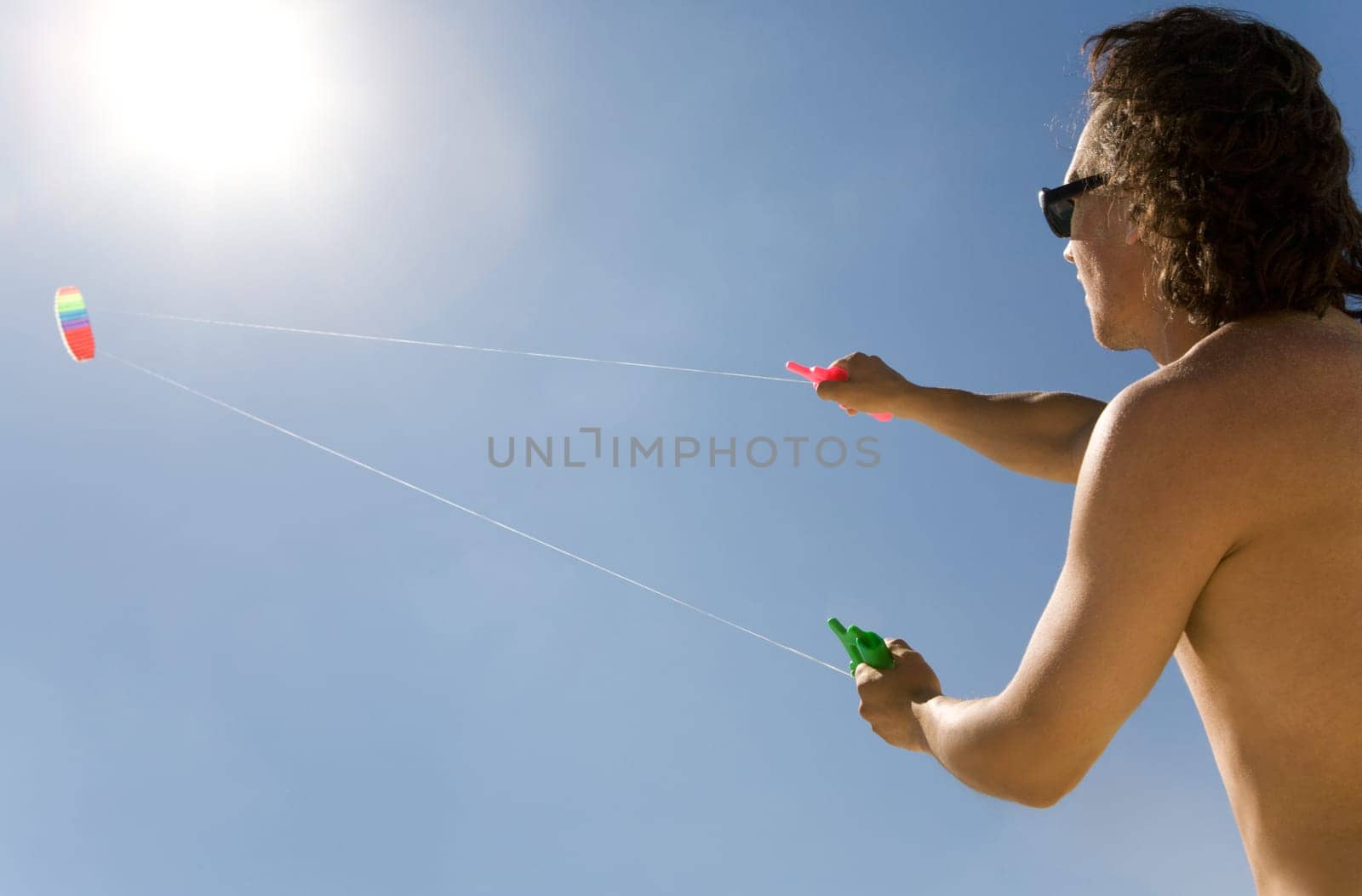 Image of young man flying a colorful kite into bright blue sky with shining sun upwards