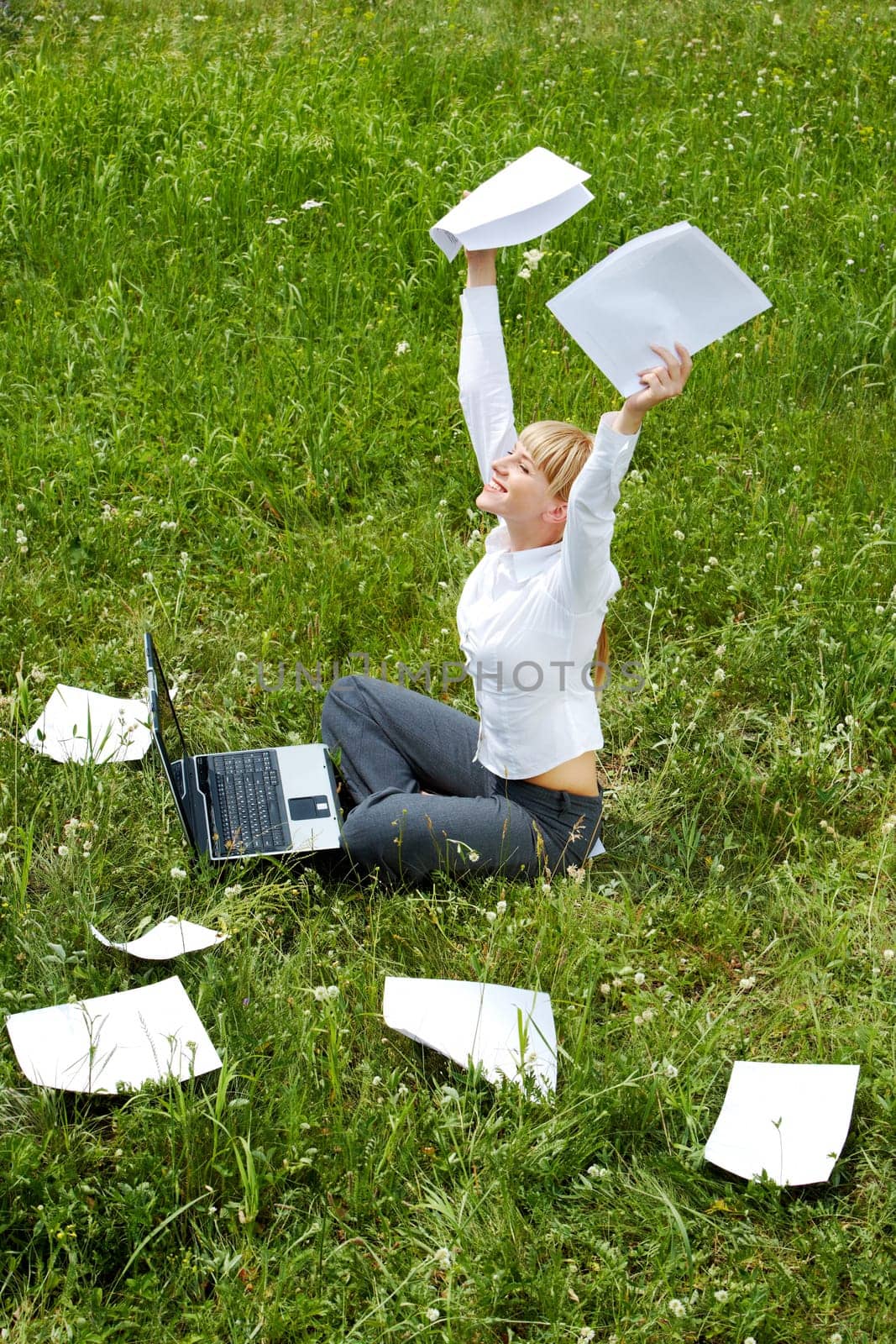 Portrait of happy woman with papers in hands sitting on green glade and raising her arms