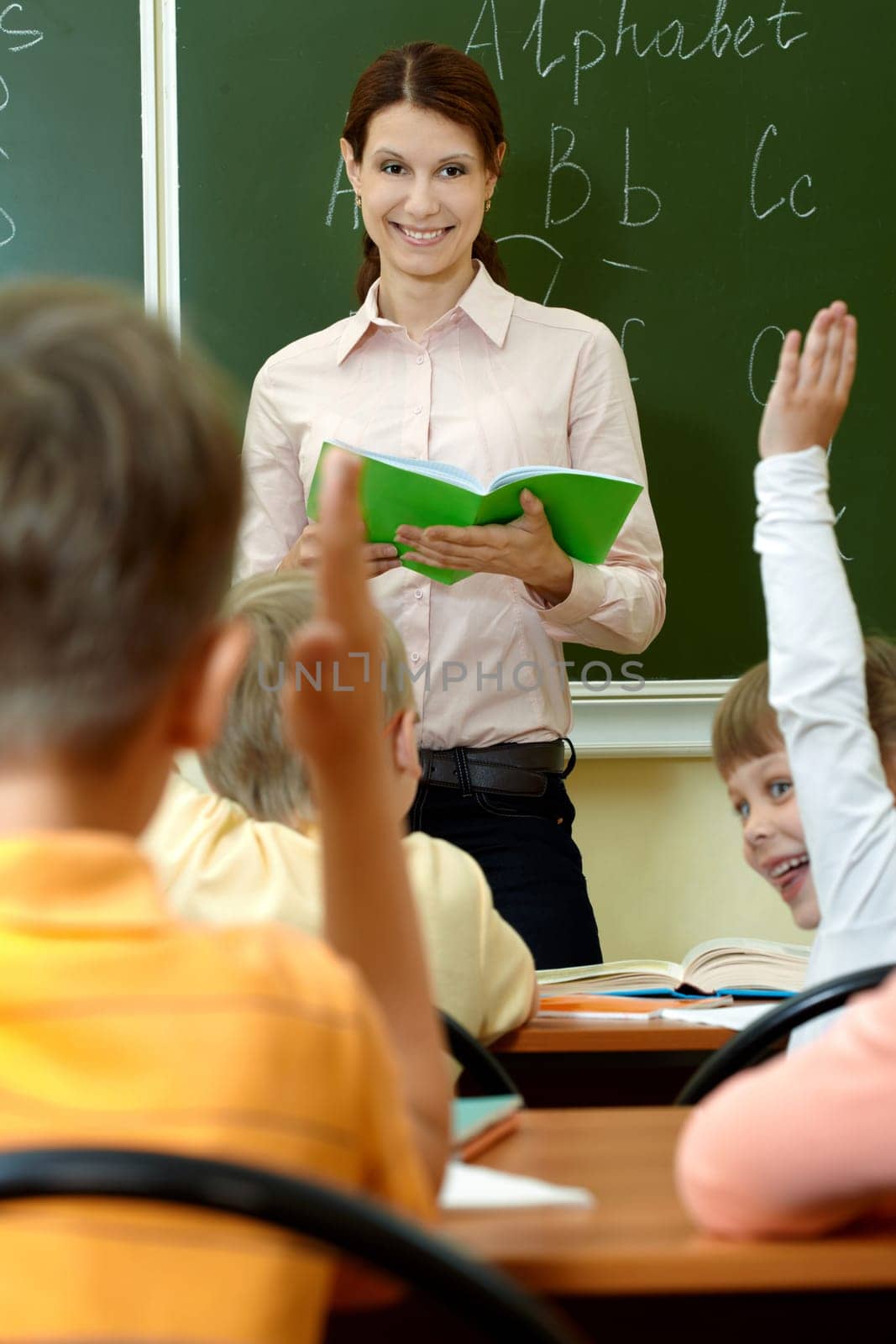 Portrait of smart teacher with exercise book looking at schoolkids in classroom