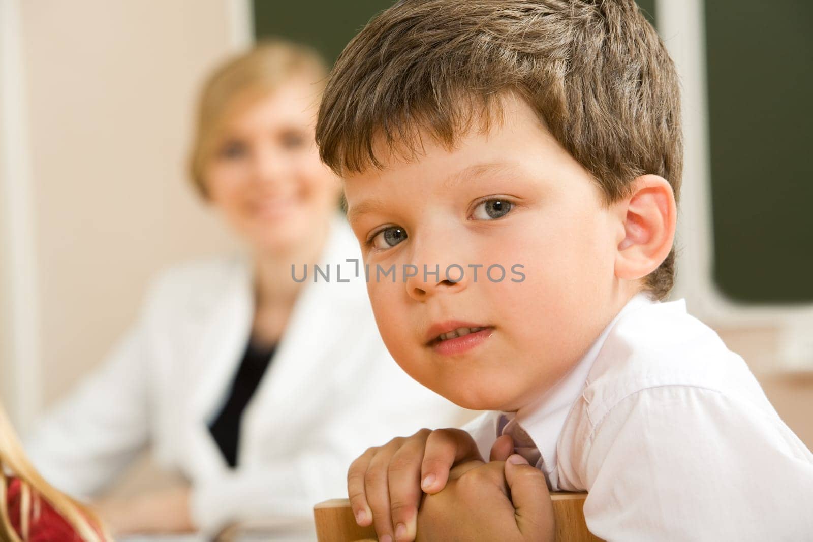 Face of clever schoolboy looking at camera in classroom with his teacher at background