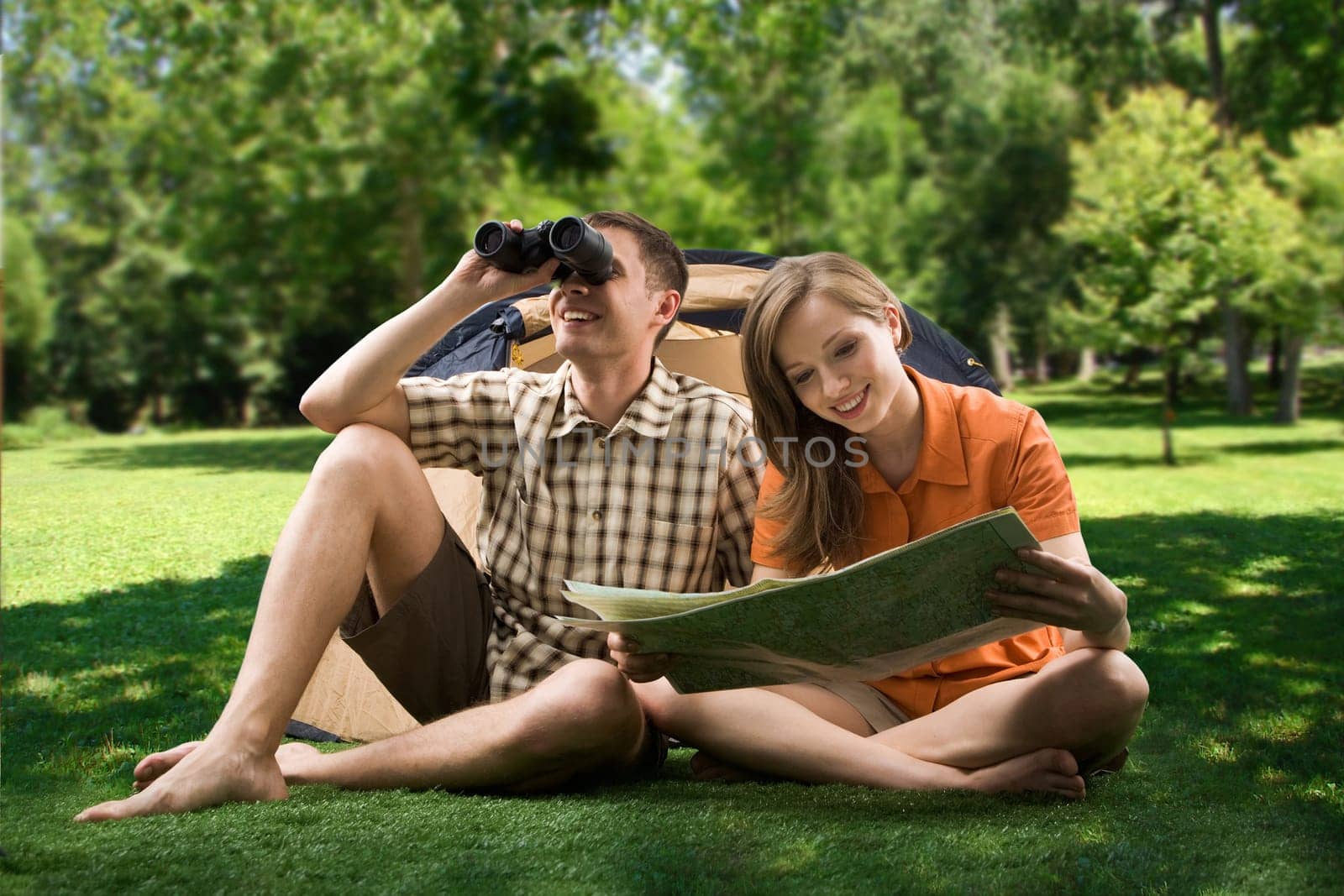 Photo of resting couple sitting on green grass while girl reading paper and guy watching through binoculars