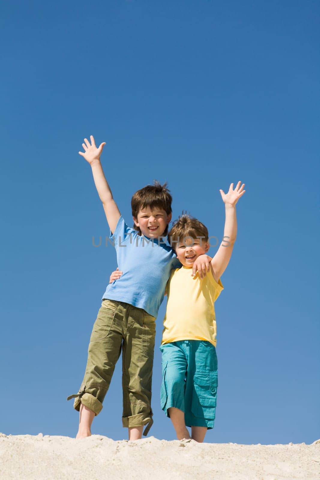 Photo of happy boys waving on sandy beach and looking at camera