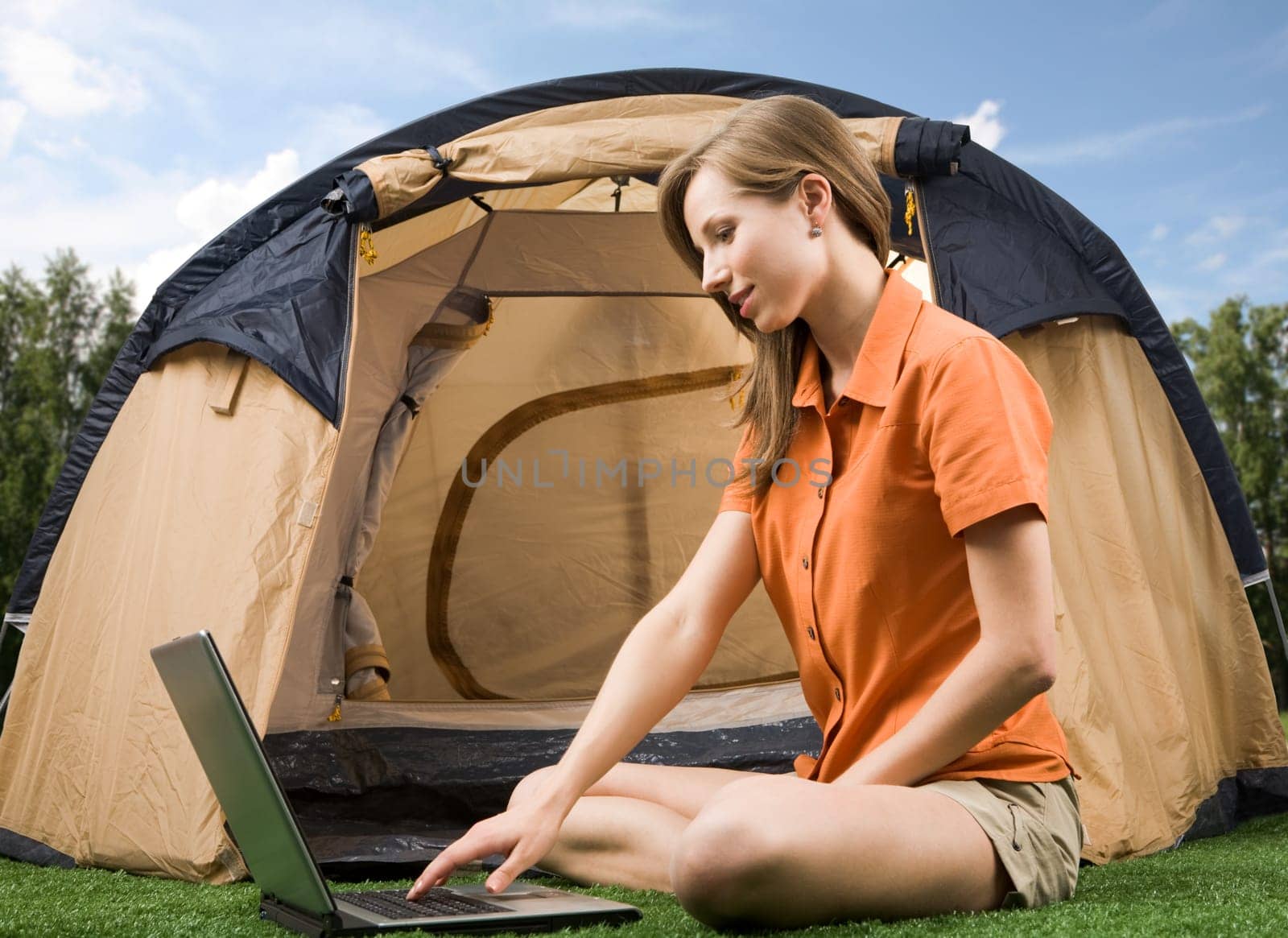 Photo of charming female sitting on green grass and typing with tent near by