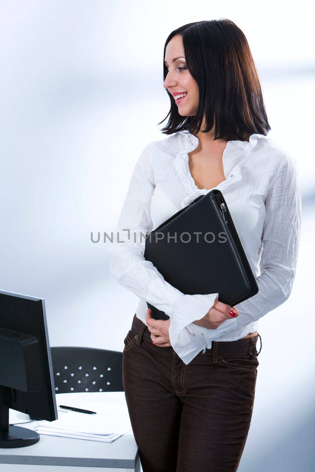 Smiling businesswoman standing near her workplace with the document case in her hands looking at the monitor