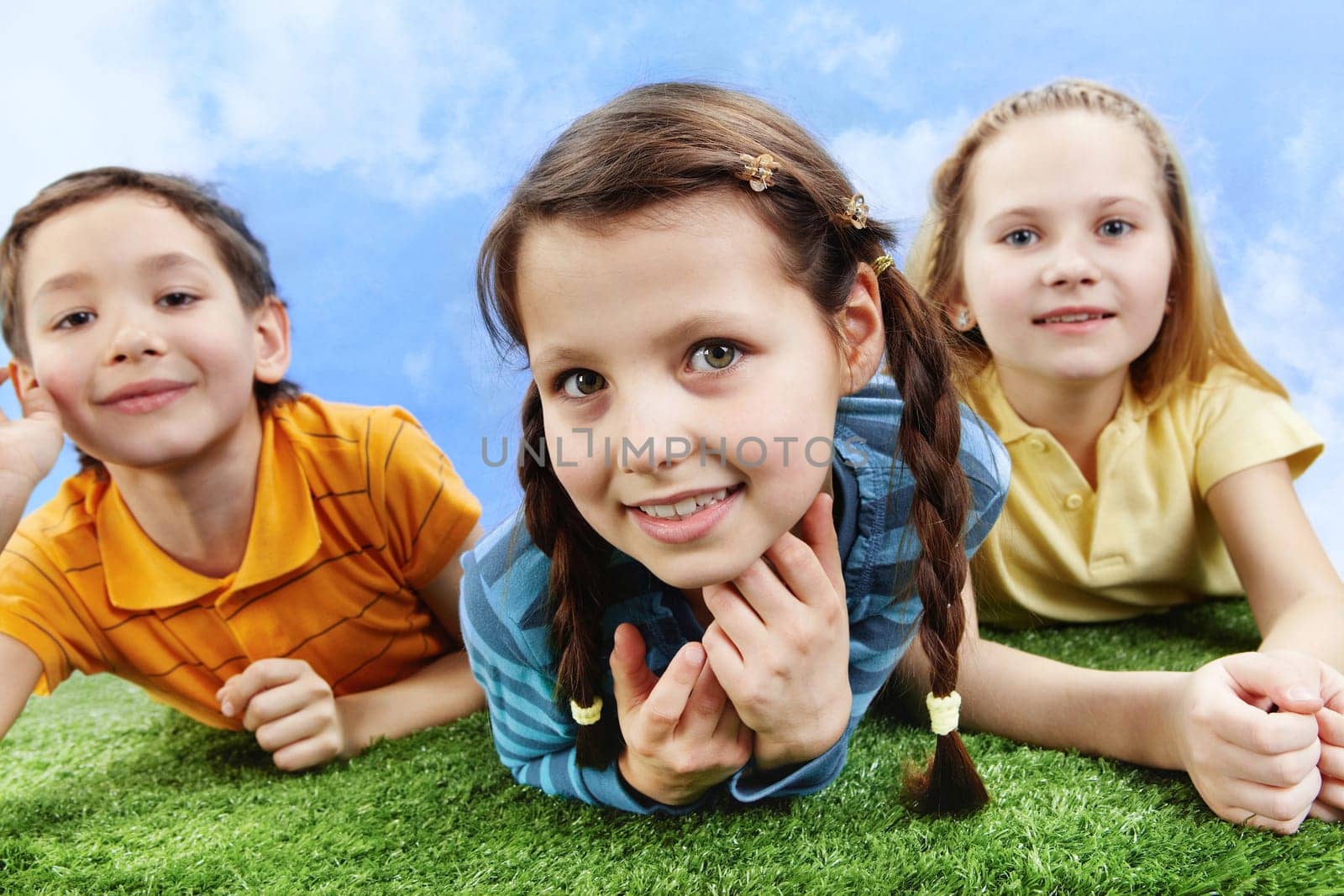 Image of happy girl lying on the grass and looking at camera with friends at background