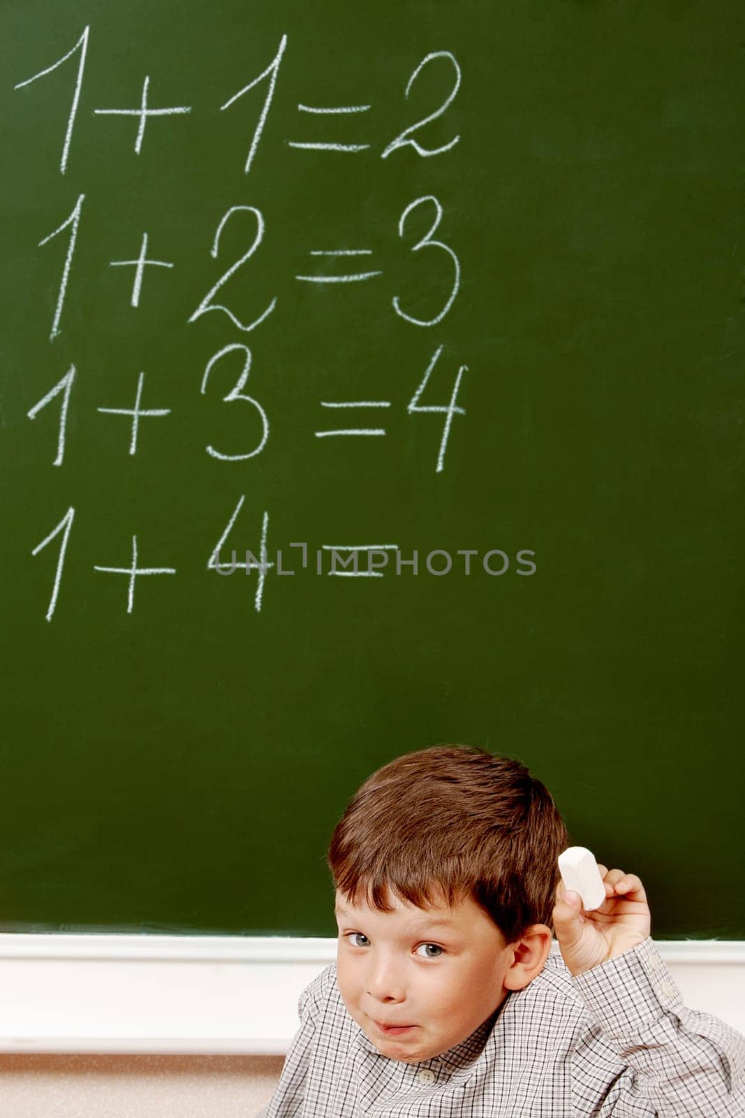 Portrait of curious schoolchild standing at blackboard with written sums on it and looking at camera