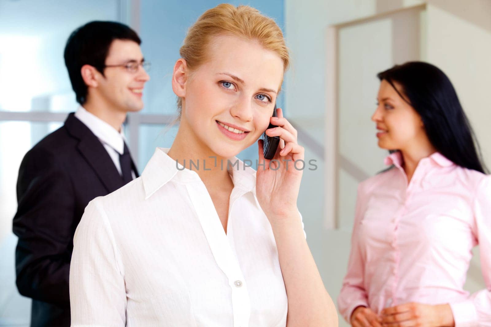 Image of pretty employee speaking on the phone and looking at camera in working environment