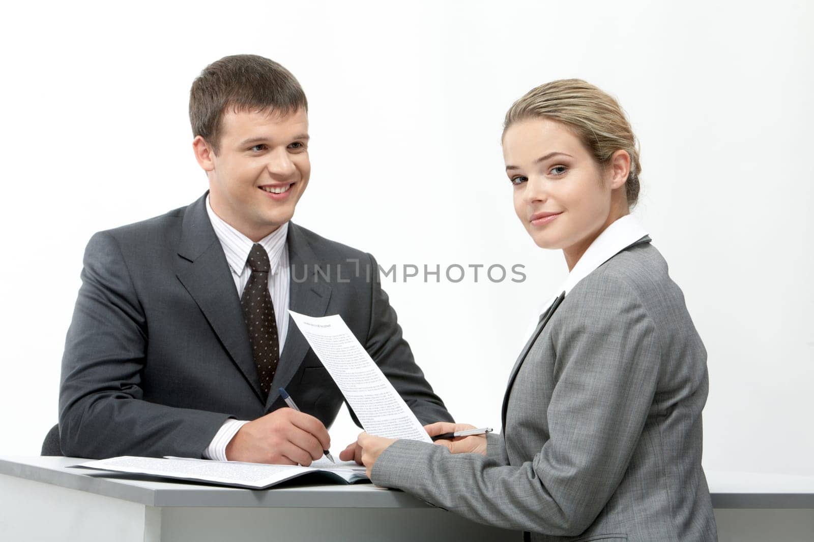 Photo of friendly secretary sitting at table and looking at camera with her partner near by
