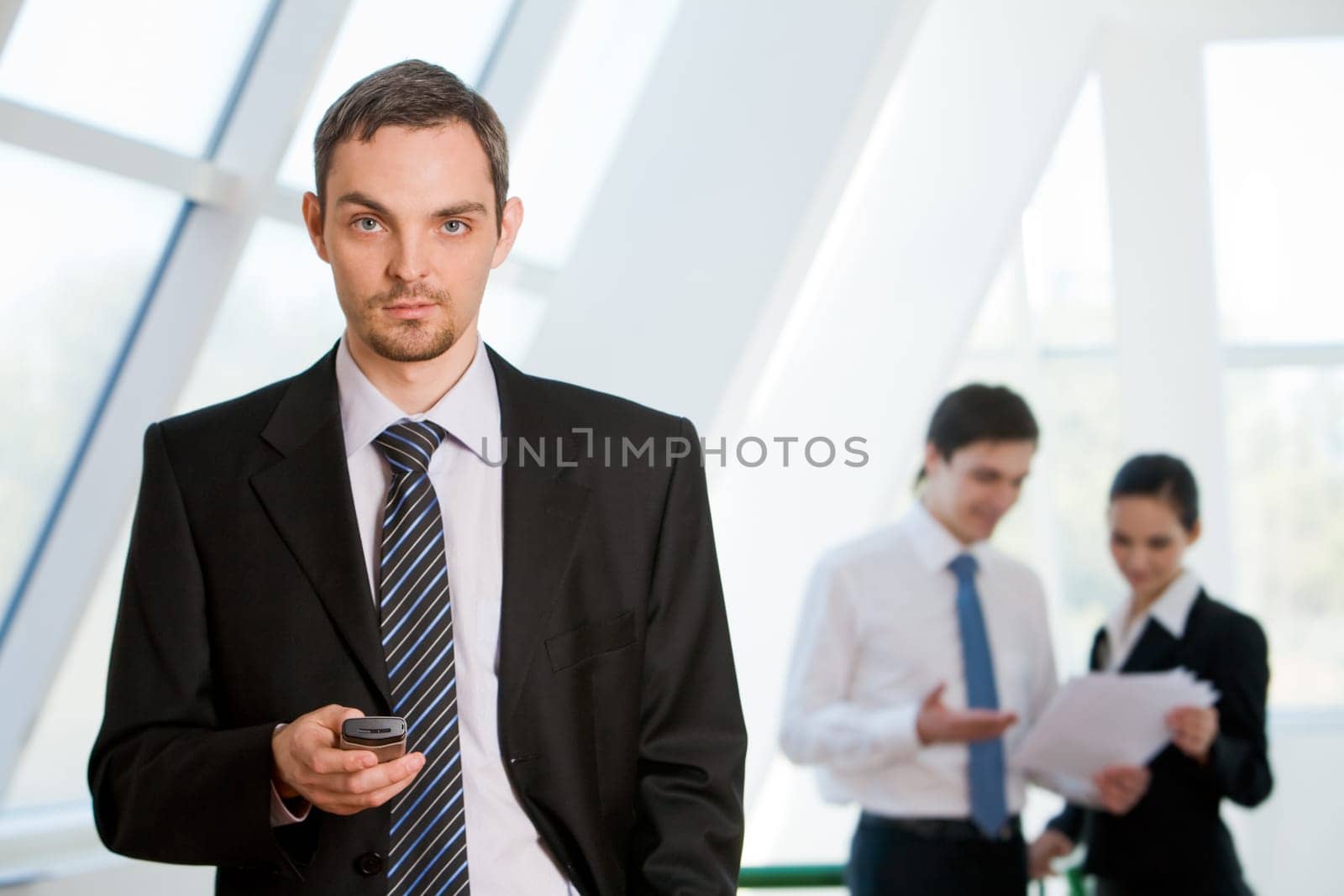 Portrait of serious businessman looking at camera with cell in hand on background of interacting co-workers
