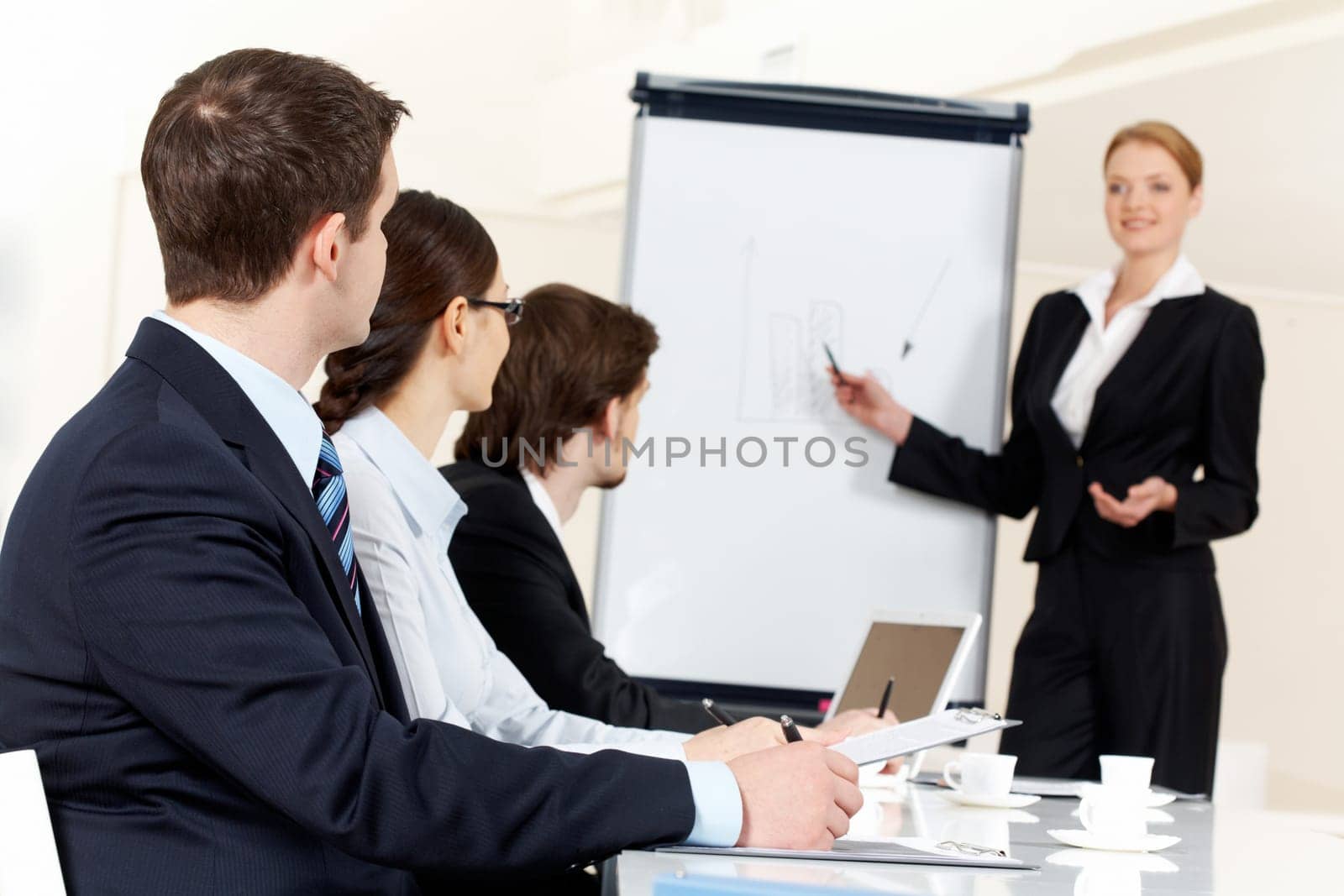 Photo of serious business people listening to female manager pointing at whiteboard while presenting new project