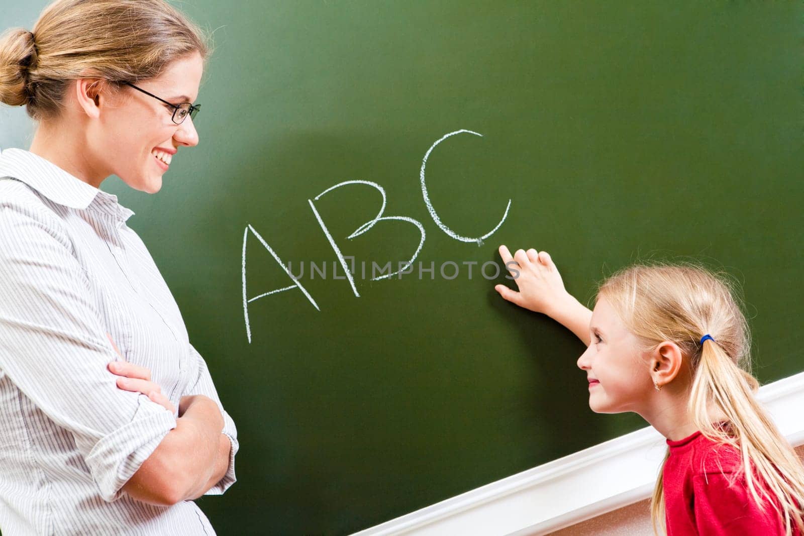 Image of smart girl pointing at letter on blackboard and looking at her teacher with smile