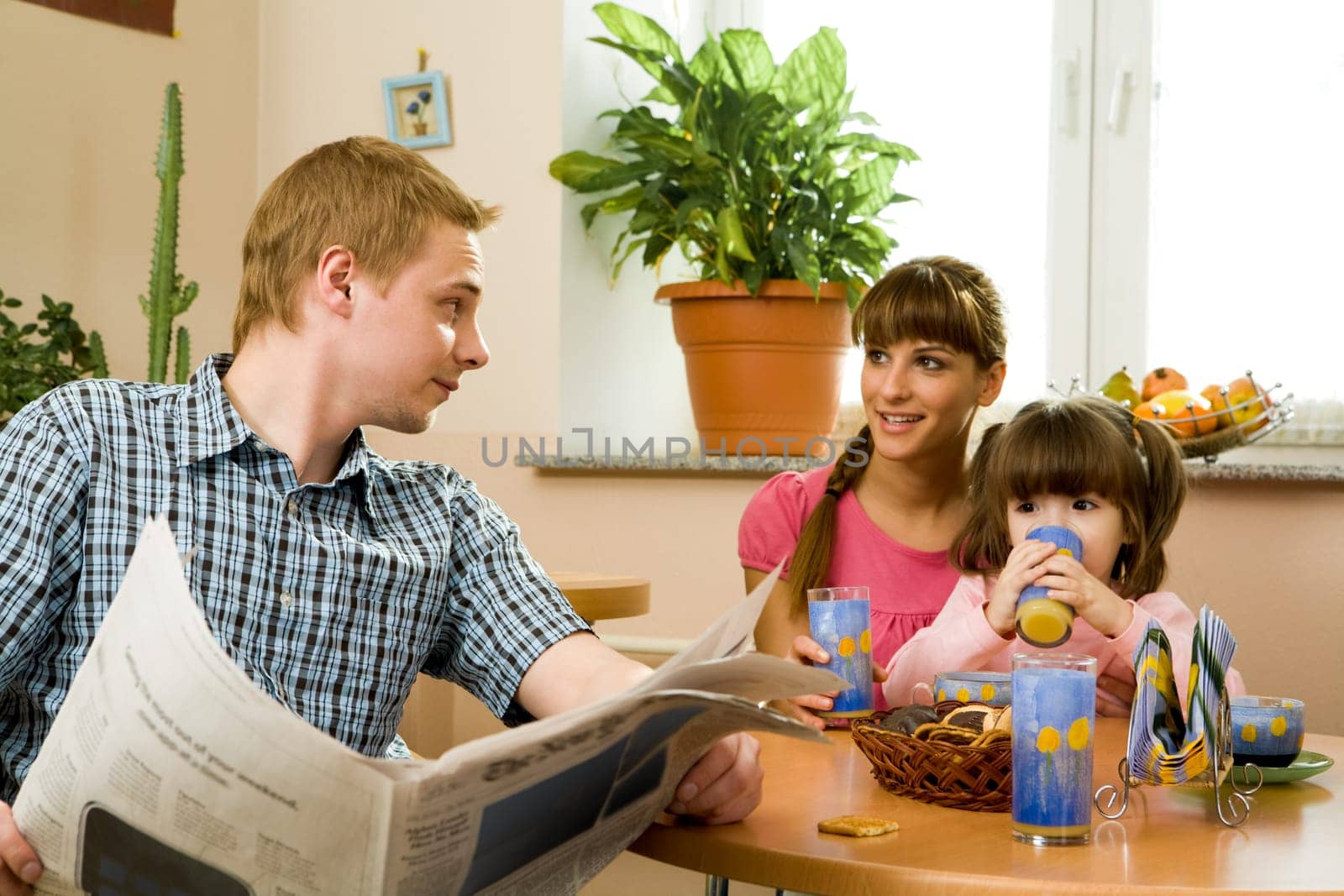 Photo of friendly woman speaking to her husband in the kitchen while he reading paper and their daughter drinking juice