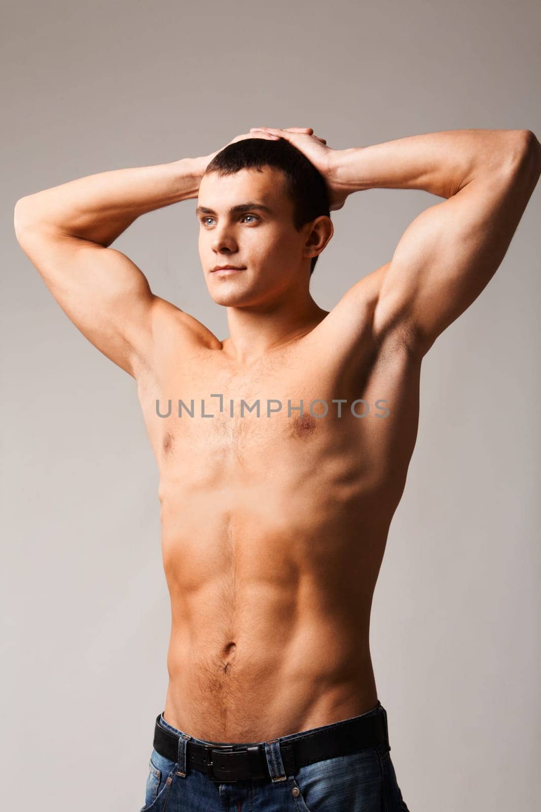 Image of shirtless man in jeans looking forwards with his hands on top of head