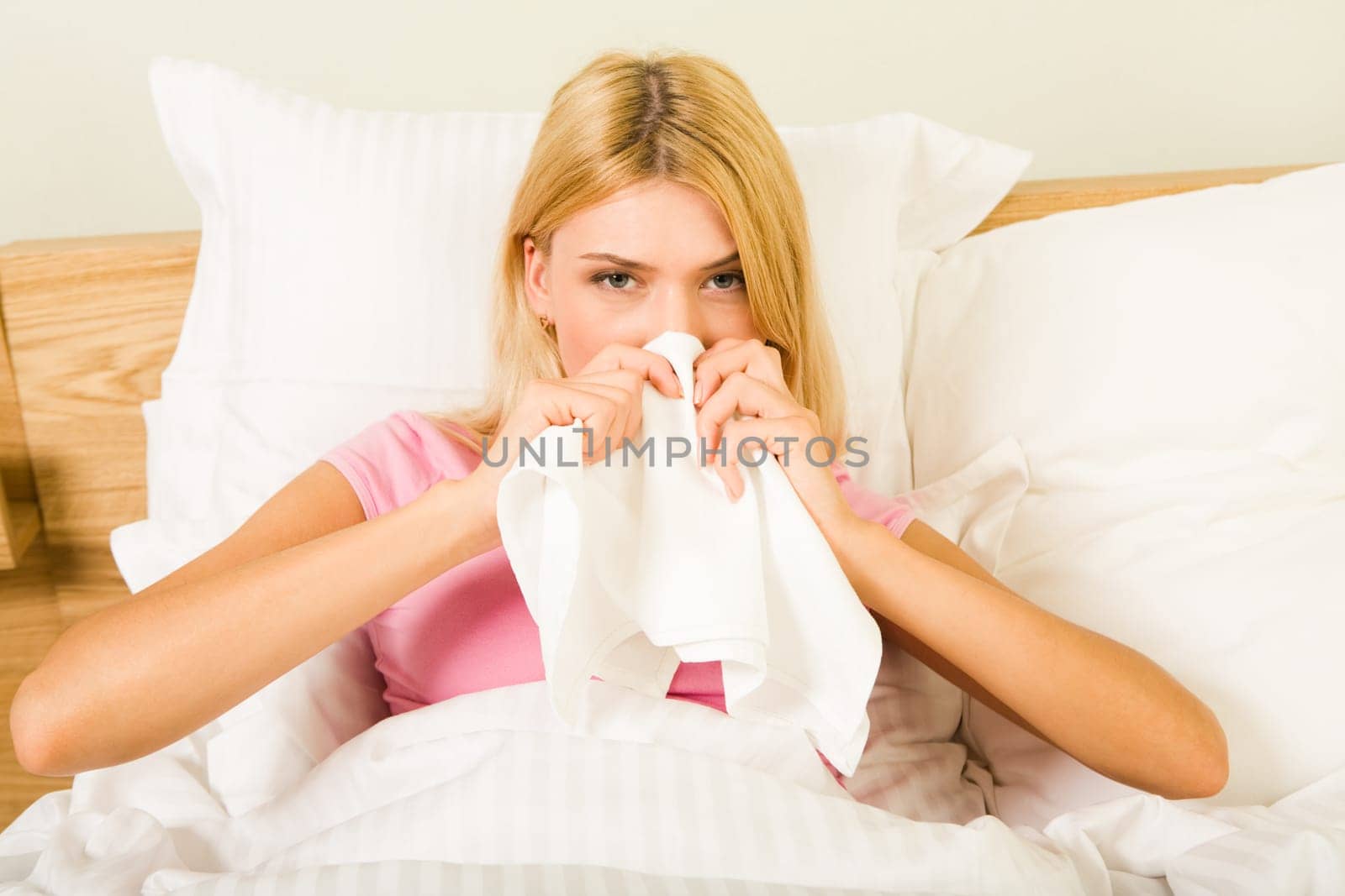 Photo of sick woman sitting in bed with tissue and blowing her nose into it