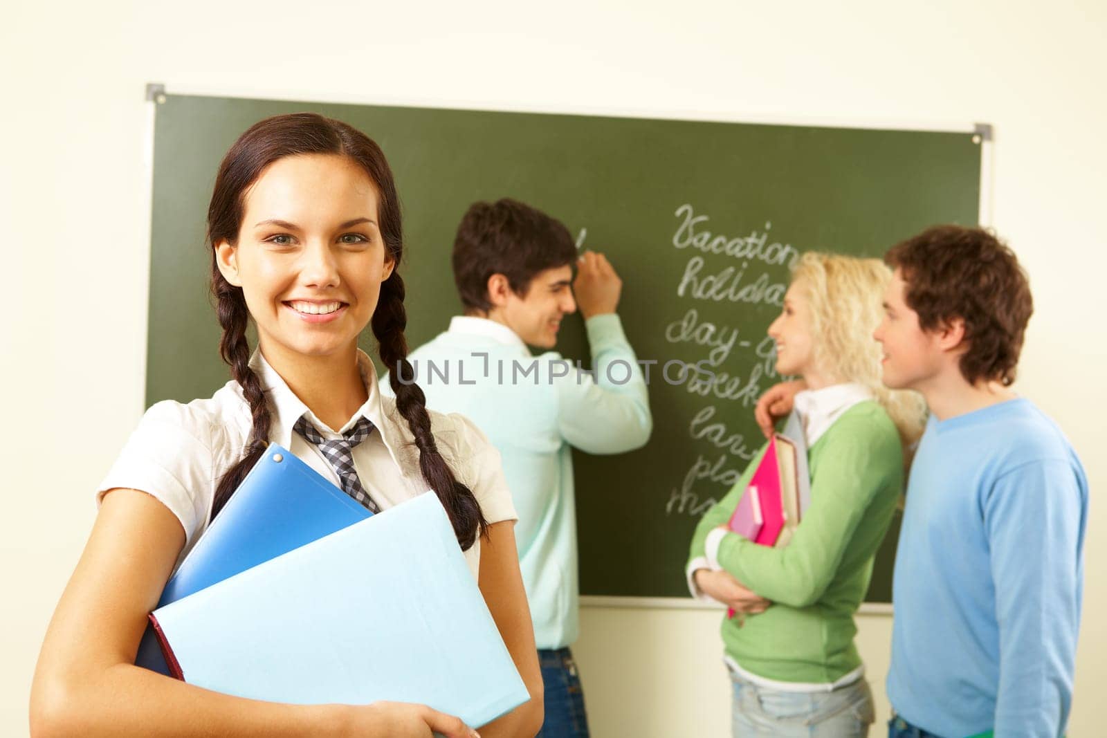 Portrait of cute girl with textbooks on background of communicating groupmates in classroom