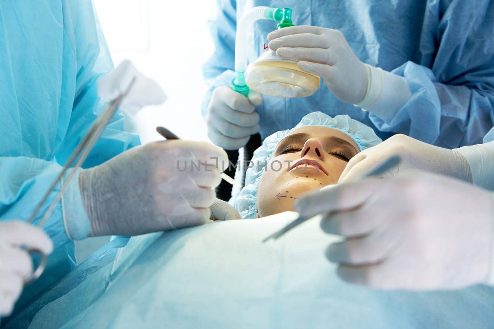 Photo of young female patient lying with closed eyes before operation with hands of doctors over her