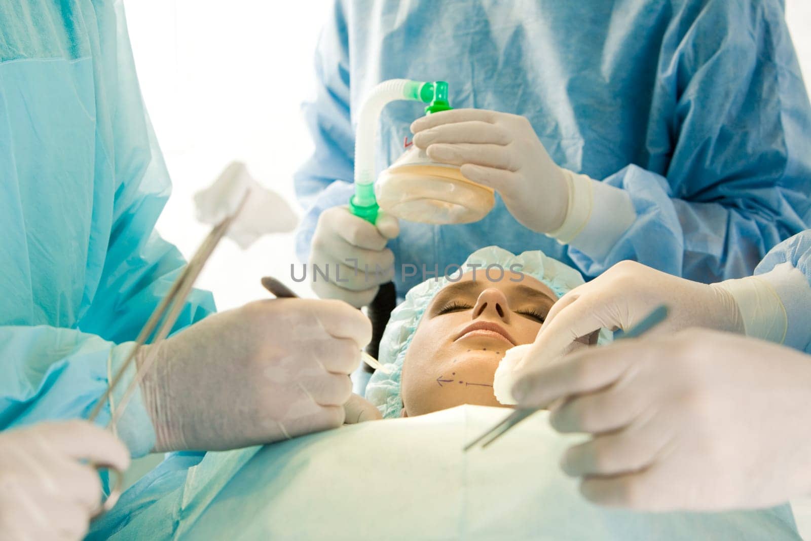 Photo of young female patient with closed eyes before operation with hands of surgeons over her holding surgical instruments and oxygen respirator