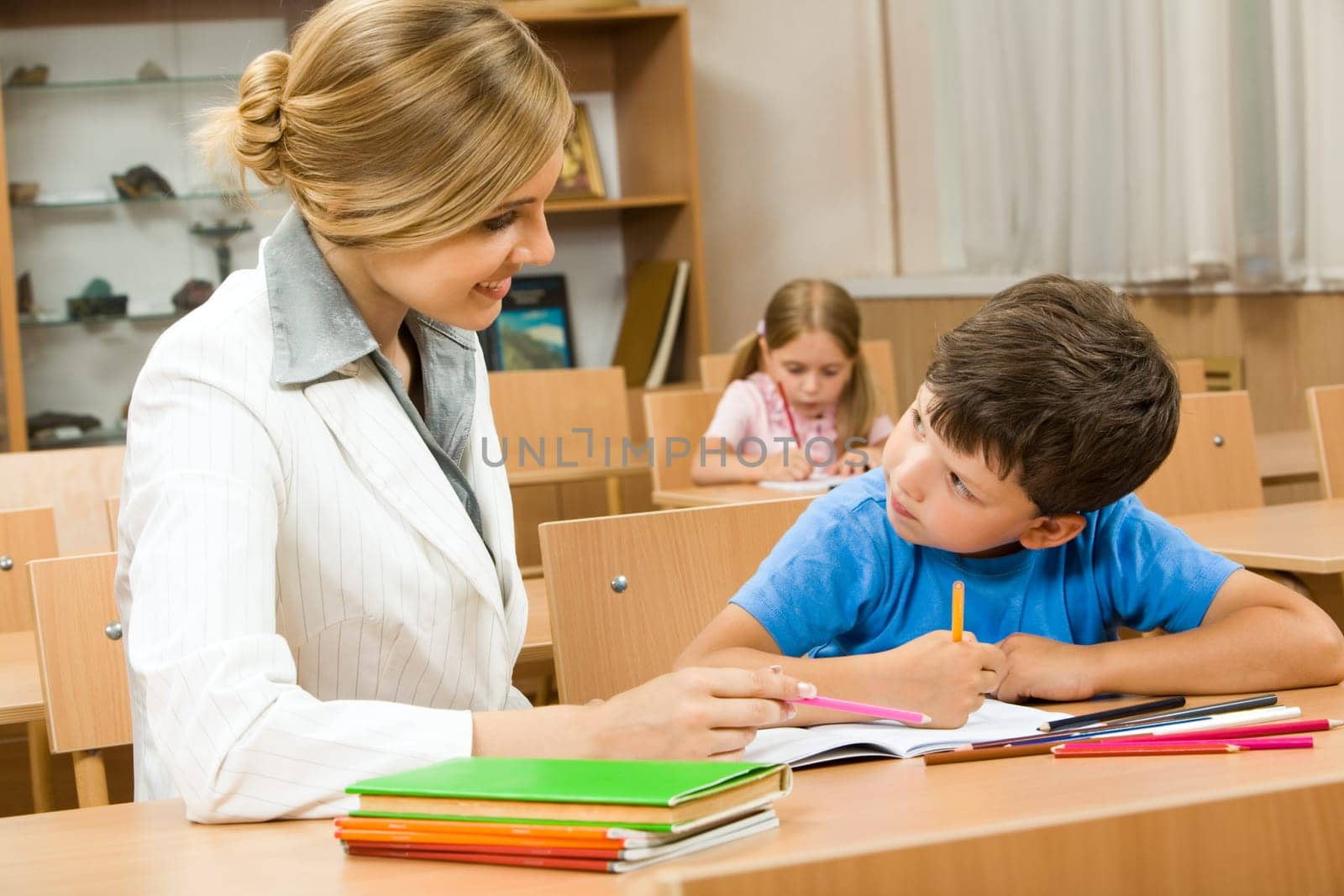 Photo of teacher and schoolboy sitting together at the desk and interacting during lesson