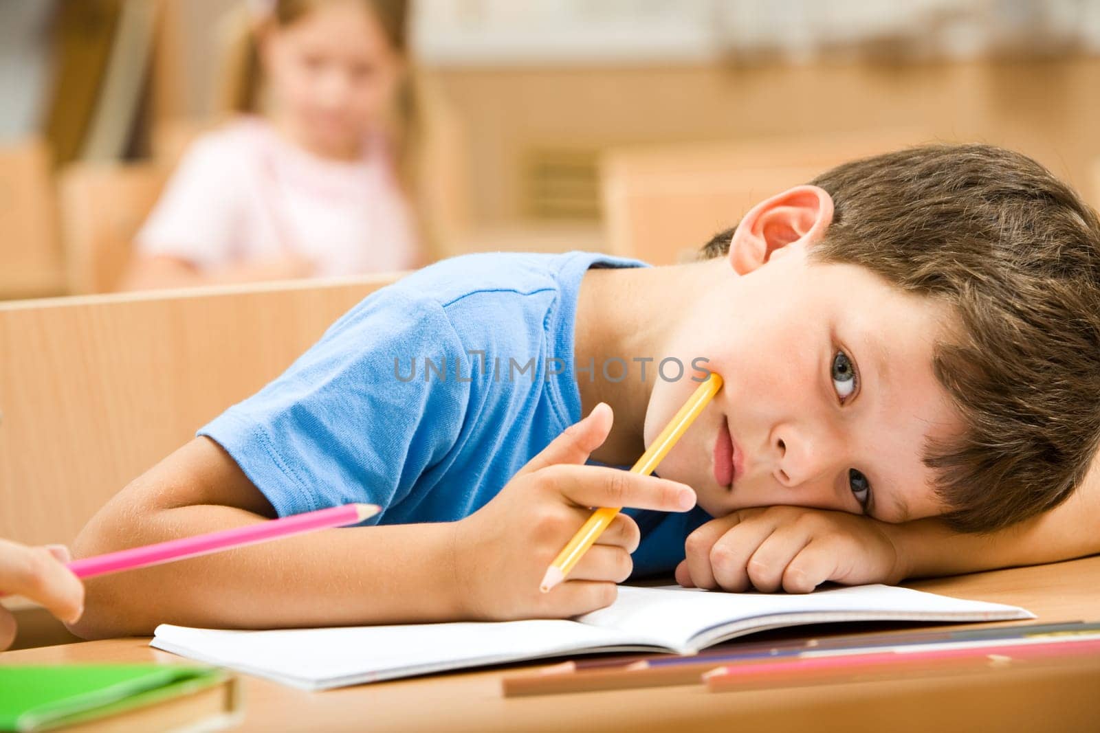 Close-up of tired pupil with yellow crayon putting his head on arm and looking at camera