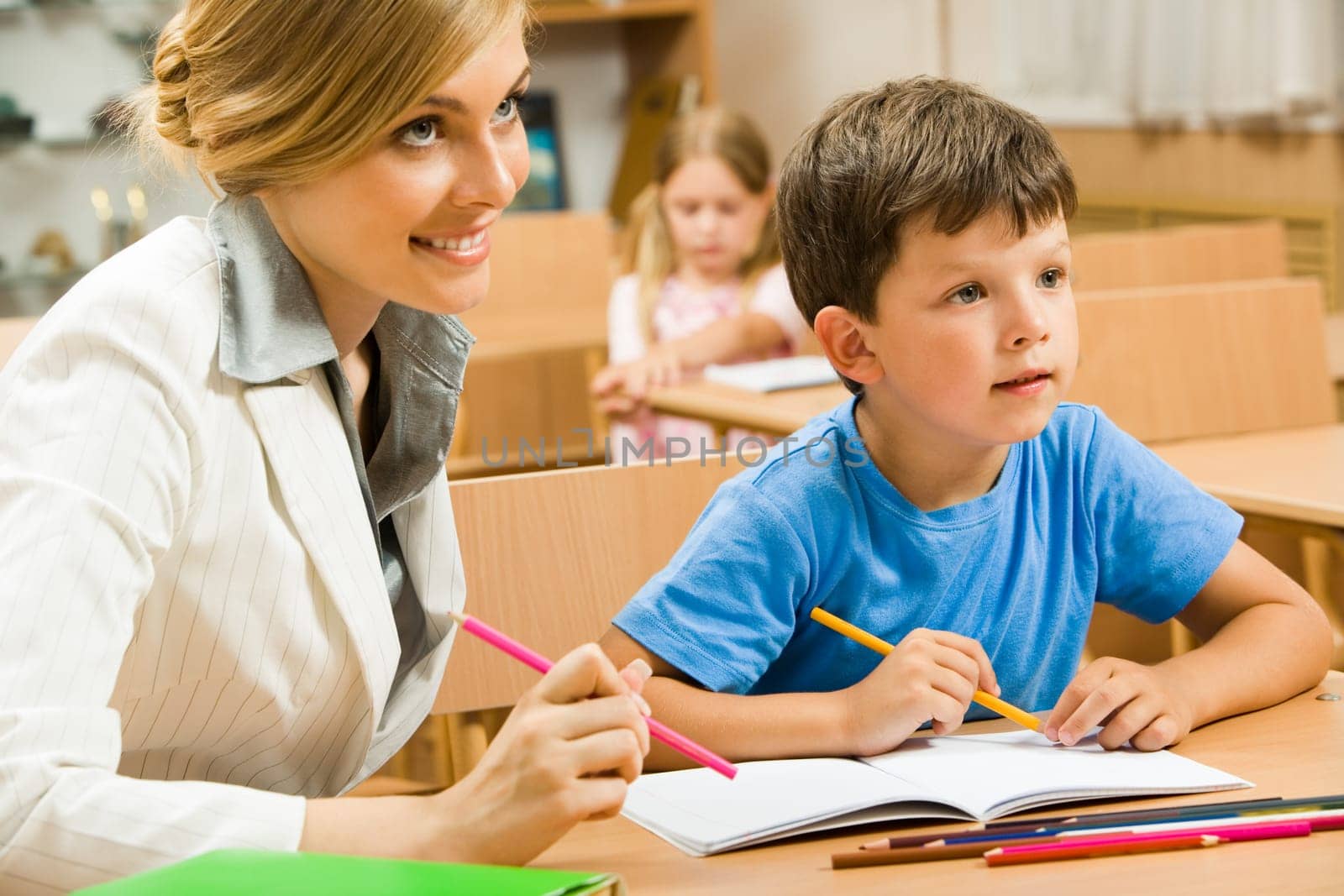 Portrait of young teacher sitting by one of pupils while both looking at something interesting