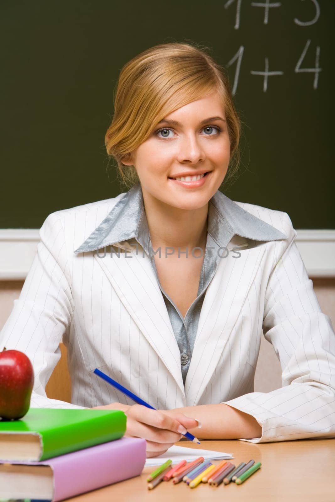 Portrait of young teacher checking up copybook with books near by and looking at camera with smile