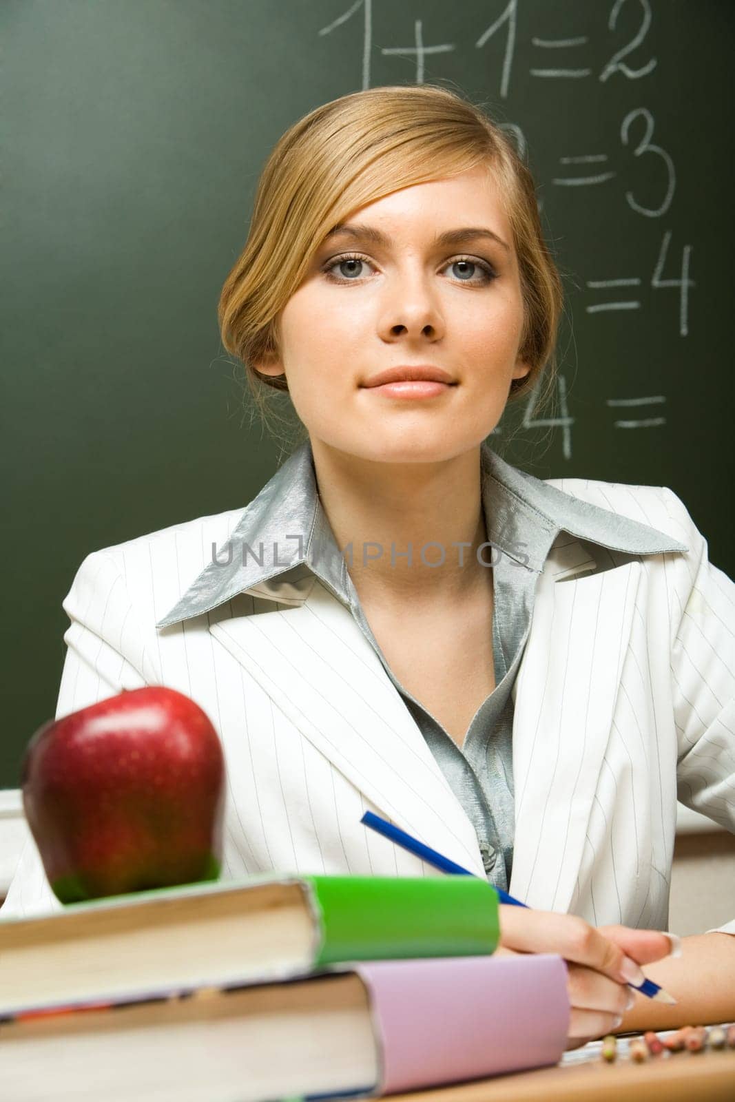 Portrait of confident teacher sitting at her desk with stack of books and apple on its top looking at camera