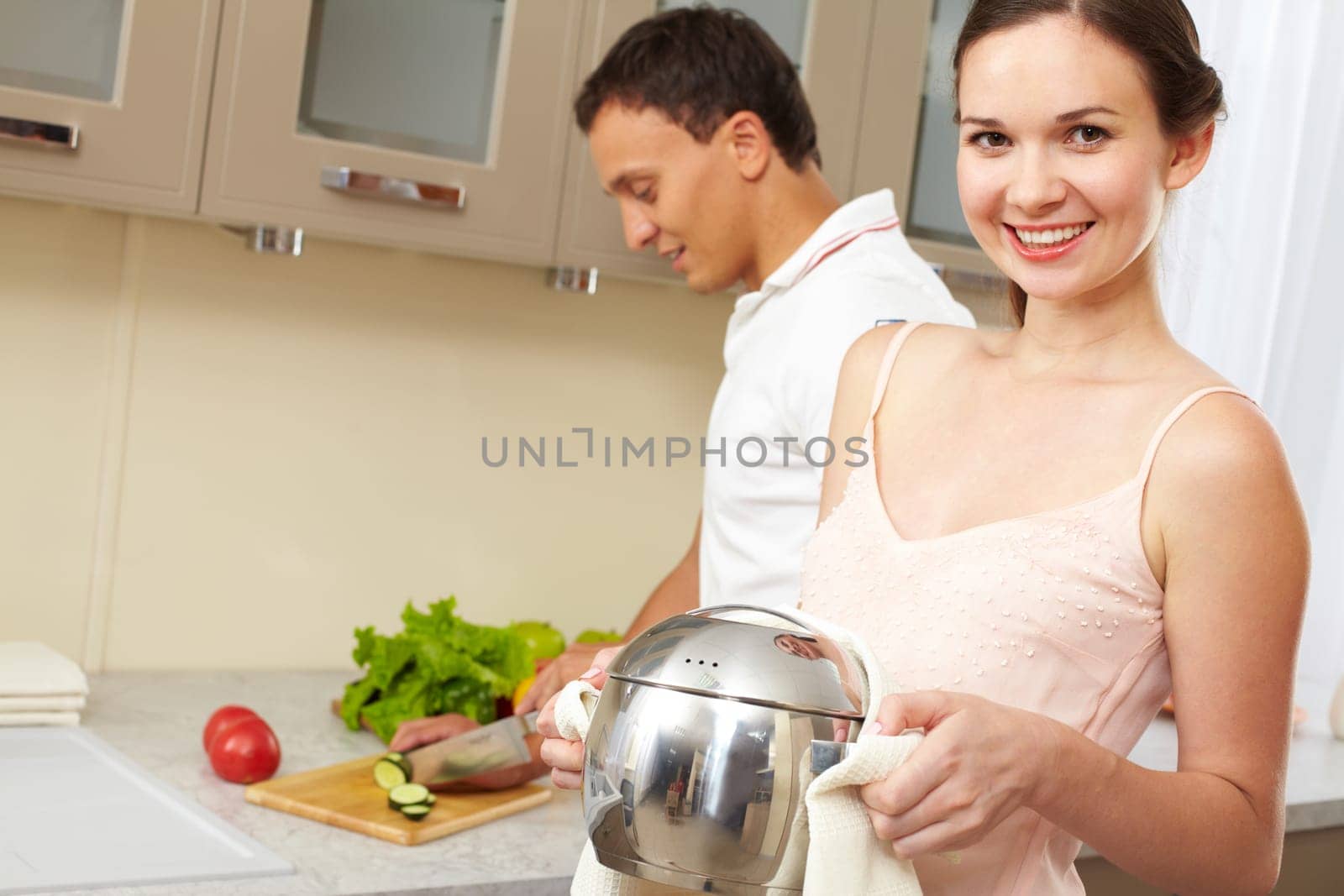Portrait of happy female with pan looking at camera while her husband cooking salad on background