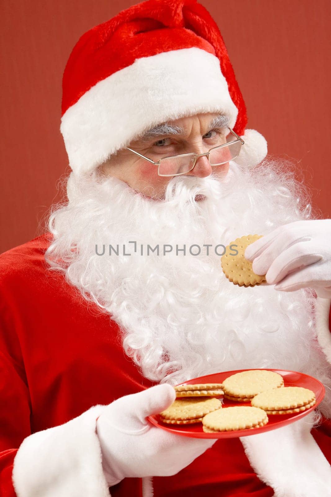 Photo of happy Santa Claus in eyeglasses looking at camera while eating biscuit
