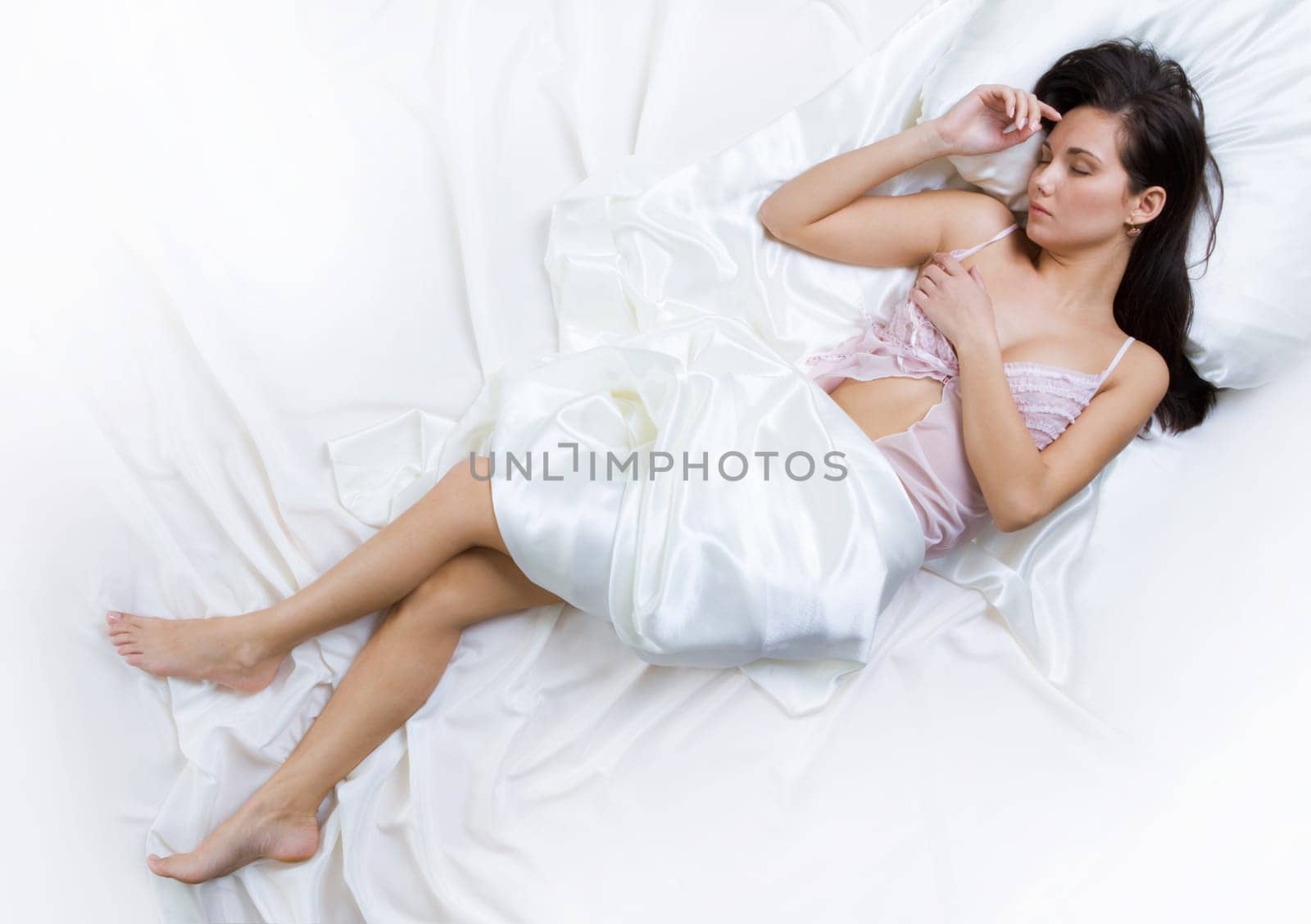 Above view of pretty female in peignoir dreaming on bed under silk sheets