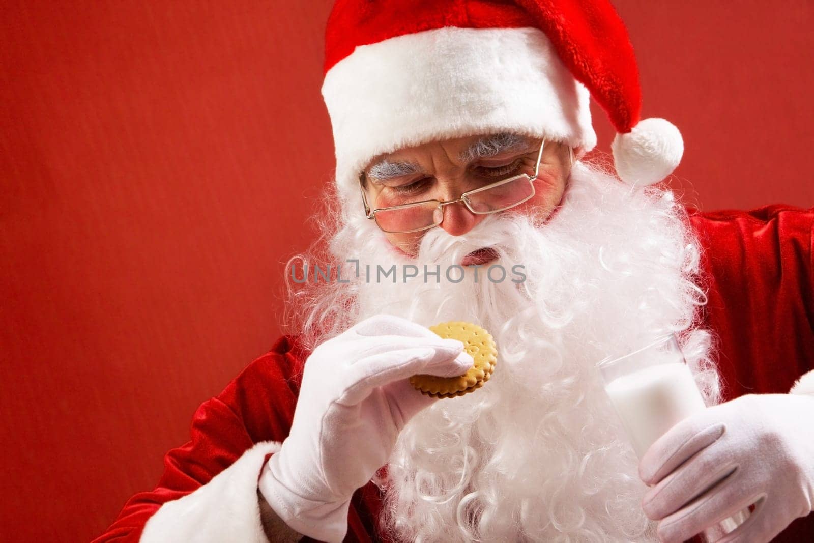 Photo of happy Santa Claus with glass of milk looking at biscuit in his hand