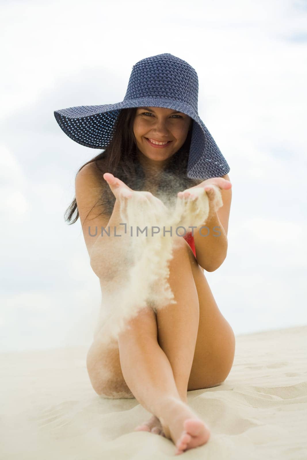 Portrait of pretty young lady in hat playing with sand and laughing at camera