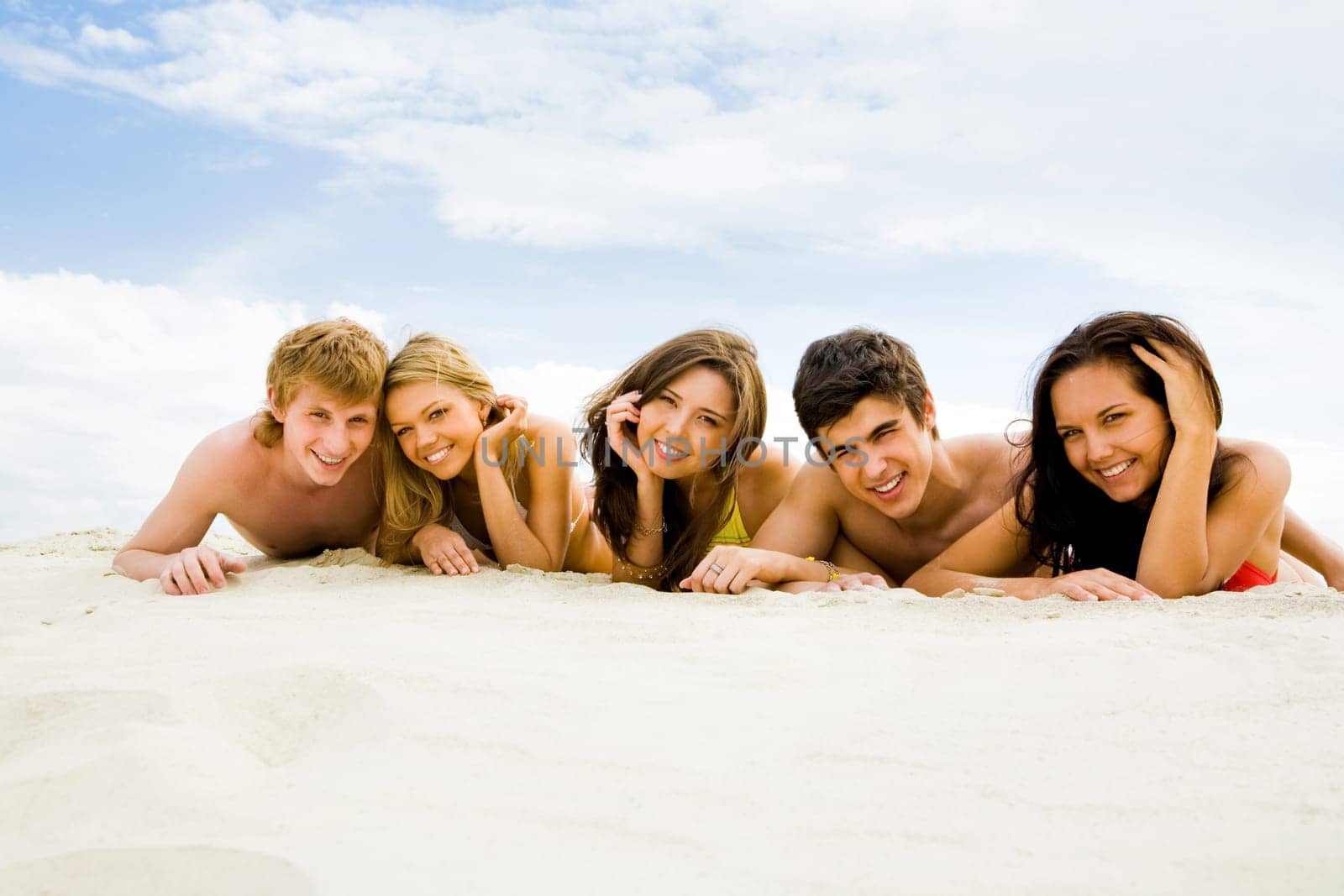 Row of cheerful friends lying on sandy beach and smiling at camera