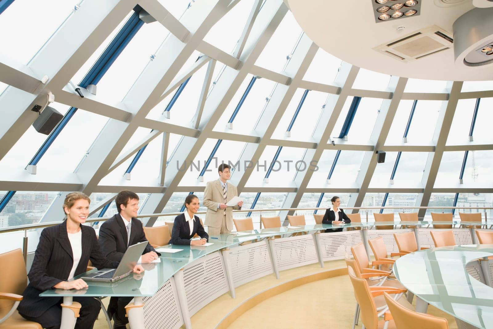Businesspeople sitting at the round table listening to their colleagues speech in large light conference hall with glassy walls discussing important questions