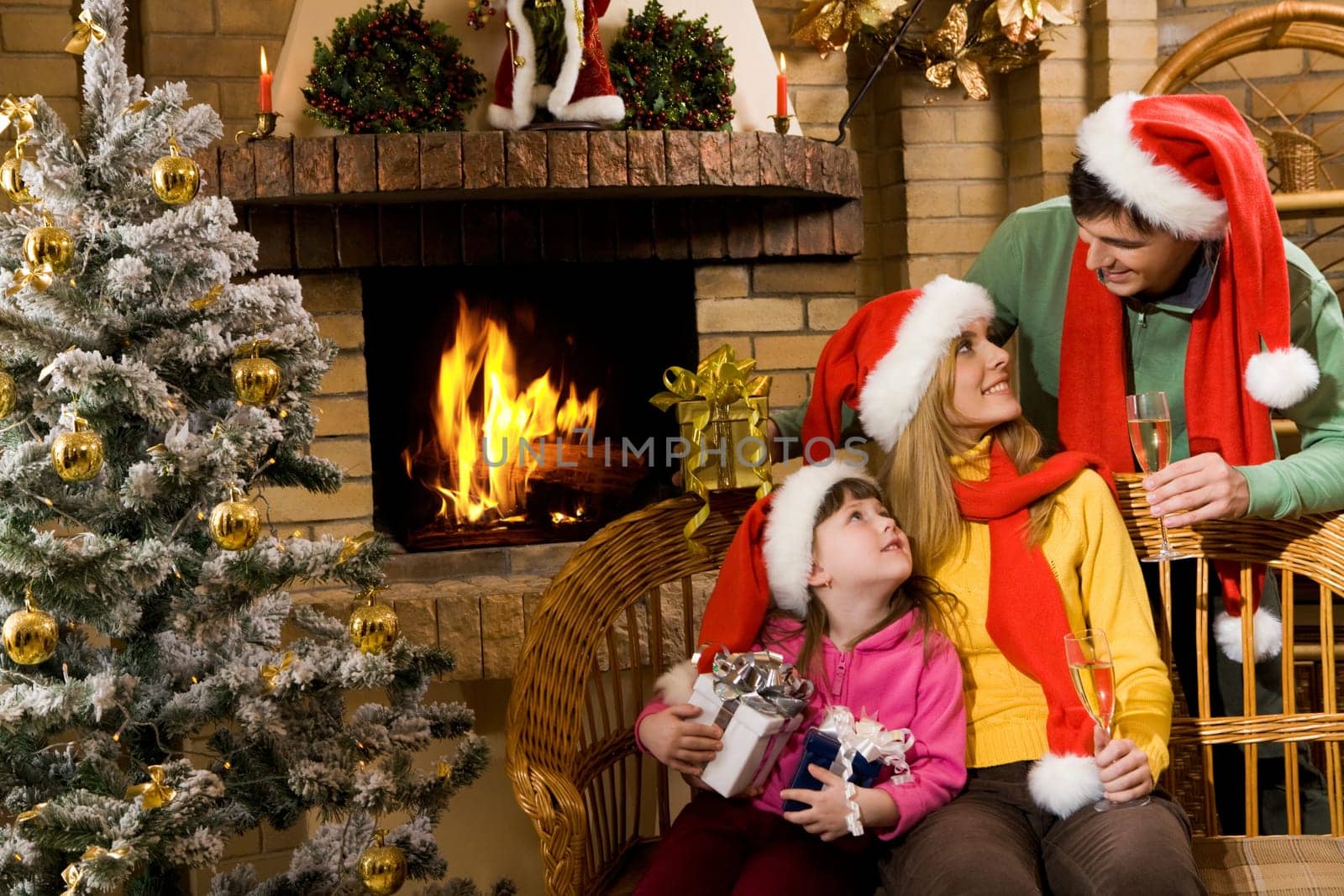 Portrait of happy family in santa caps sitting by fireplace and looking at each other