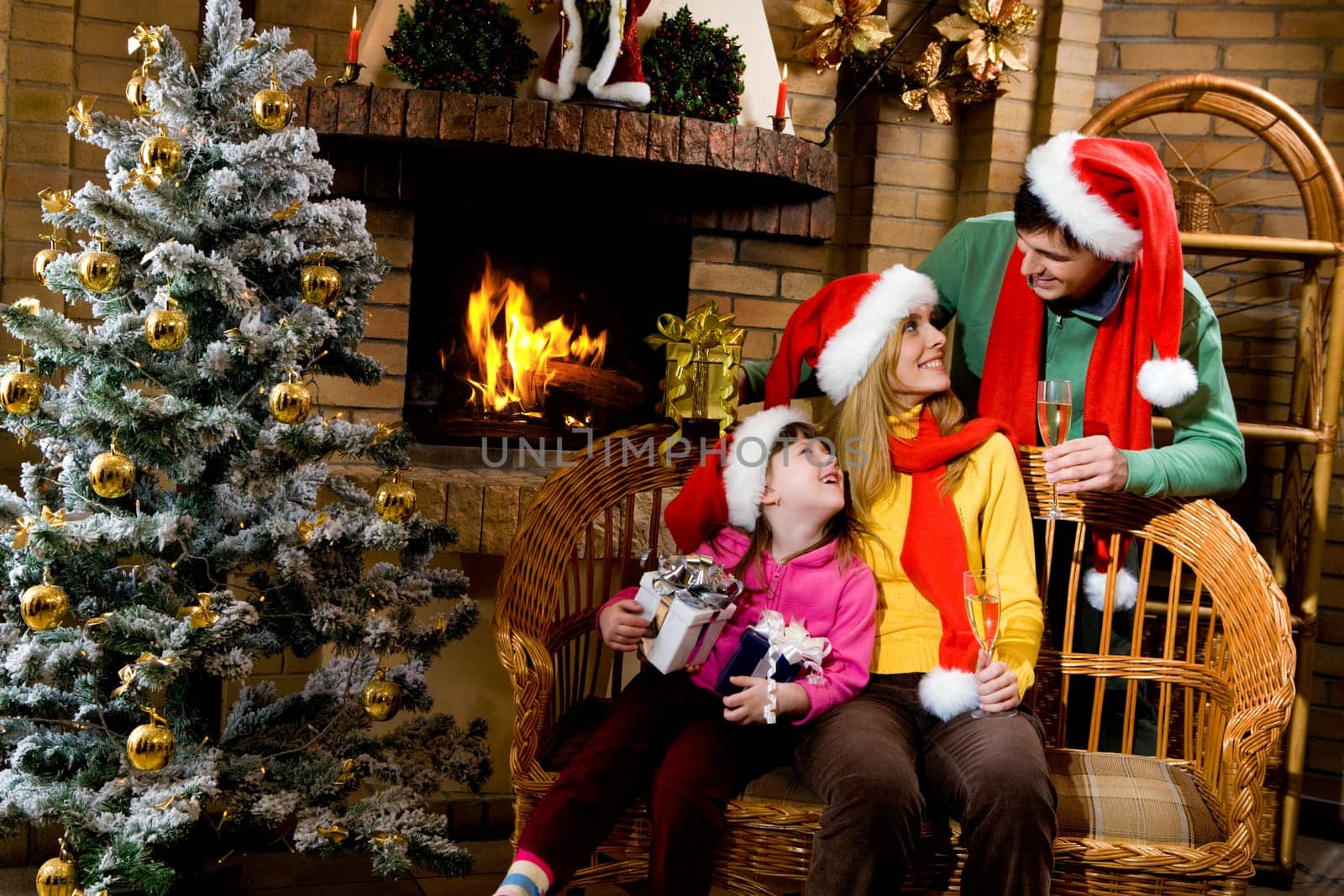 Portrait of happy family wearing santa caps while sitting by fireplace and looking at each other