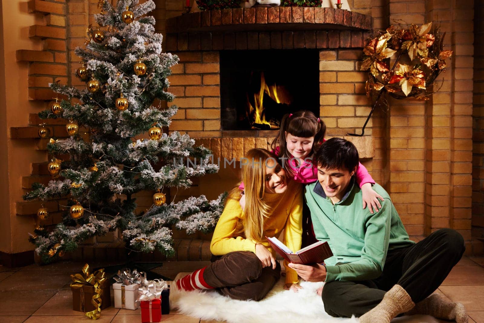Portrait of cheerful family sitting on white fur reading old book with decorated fir tree near by