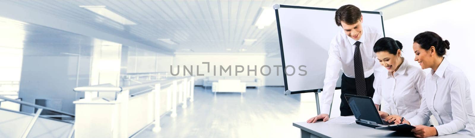Two smiling business ladies sitting at table and looking at laptop monitor while their partner standing near by