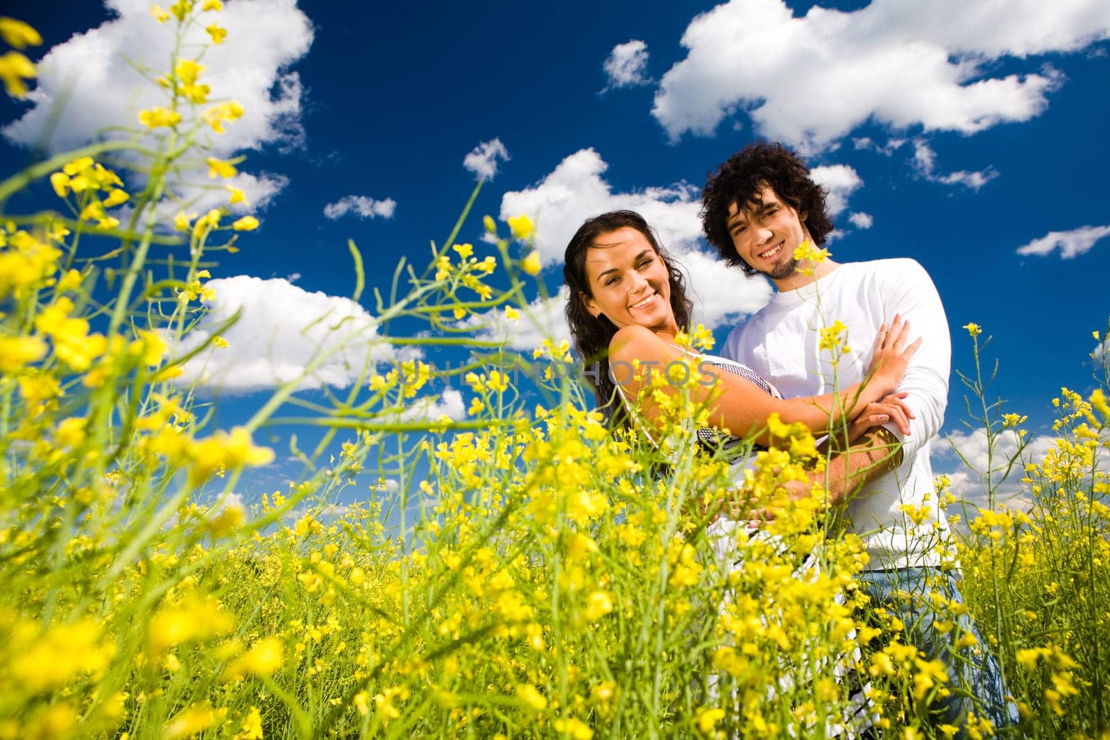 Image of smiling guy embracing his girlfriend while both looking at camera among yellow flowers