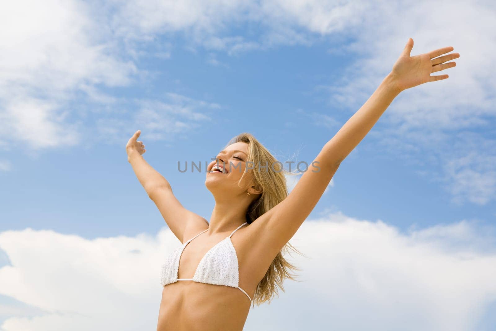 Portrait of happy girl in white bikini praising God with her eyes shut and raised arms on background of cloudy sky
