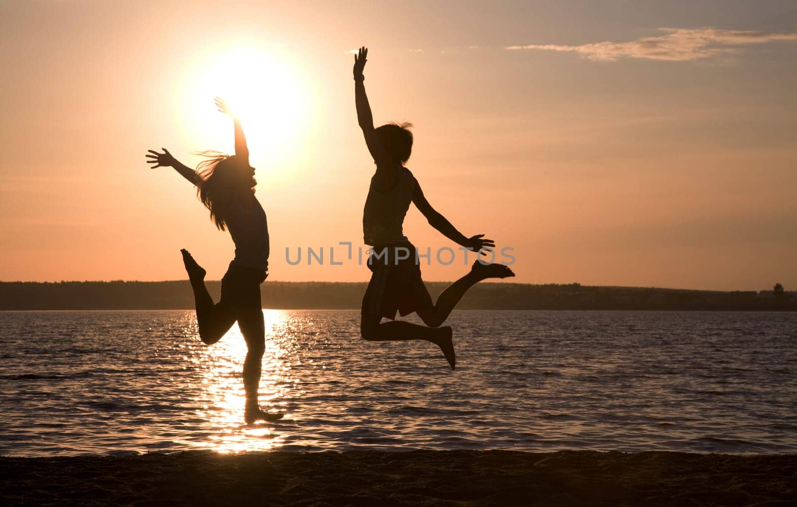 Silhouettes of happy couple jumping on background of lake at sunset