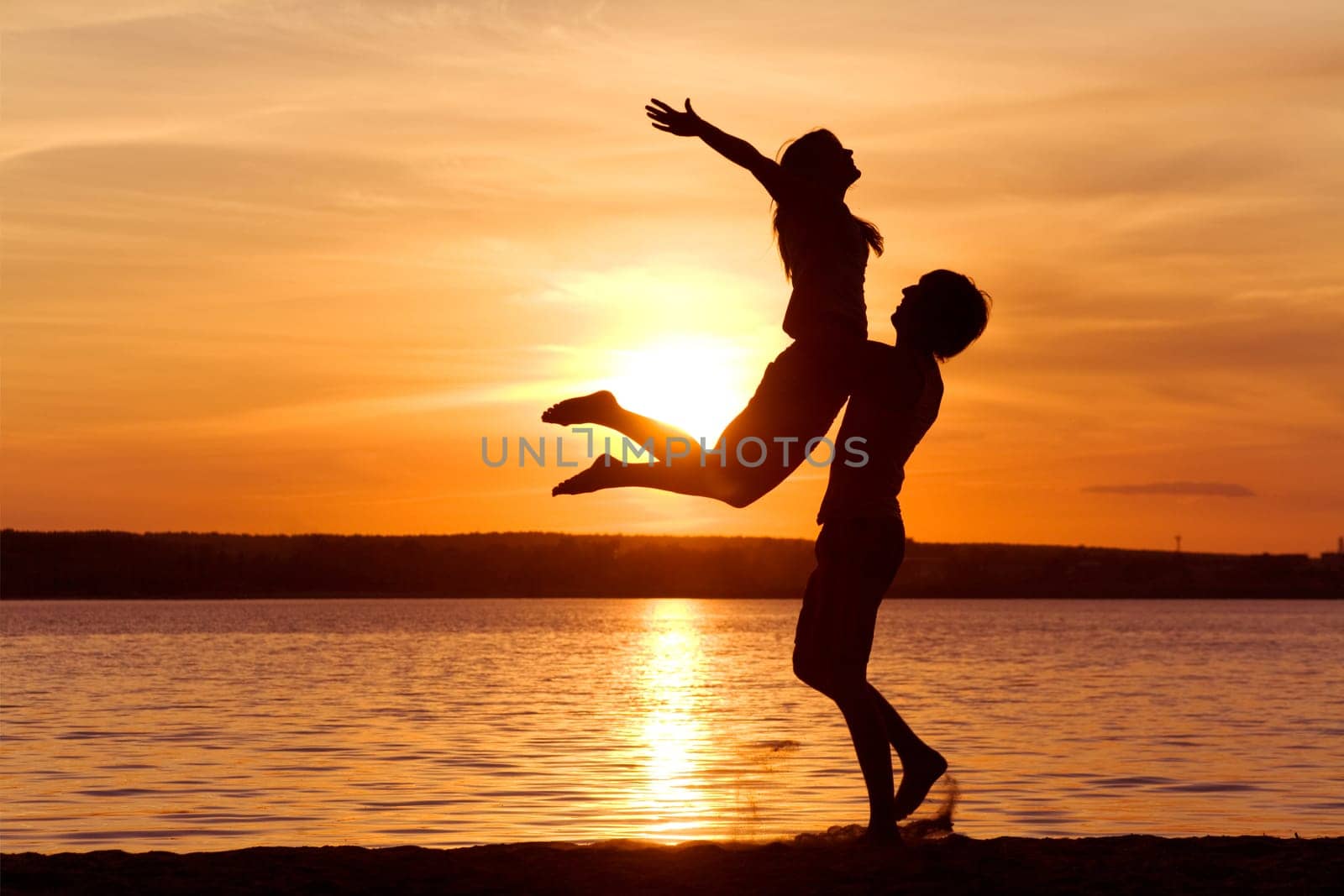 Figures of happy guy holding his girlfriend while the latter raising her arms at sunset