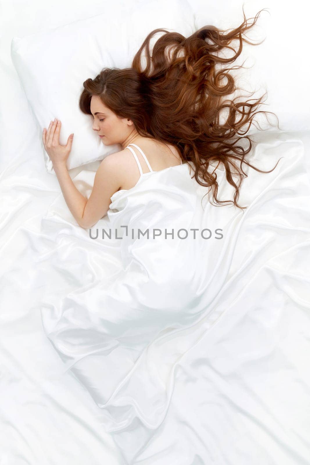 Above view of young beautiful woman sleeping in bed covered with white silky sheet