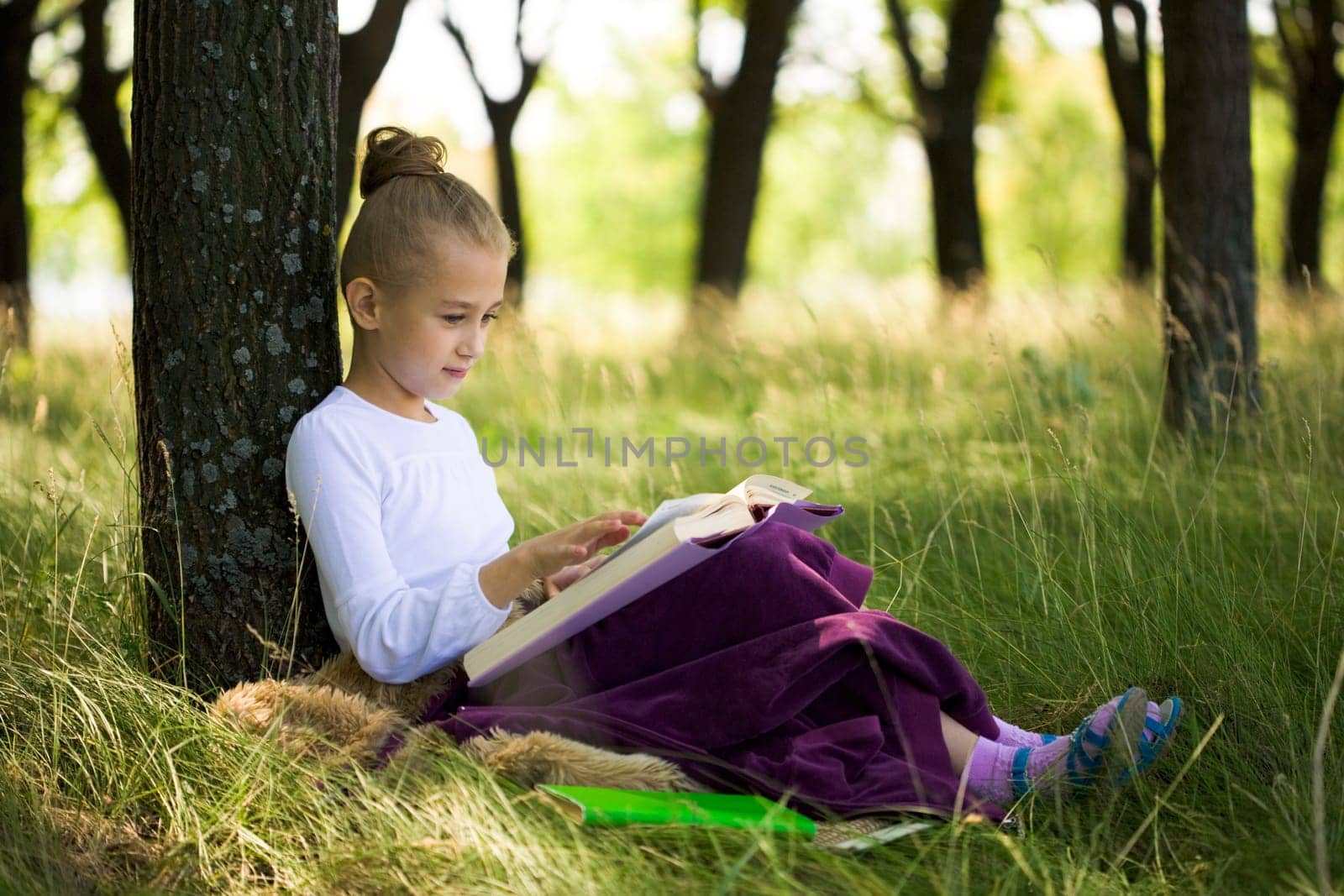 Pretty girl sitting under tree and reading at summer in a park
