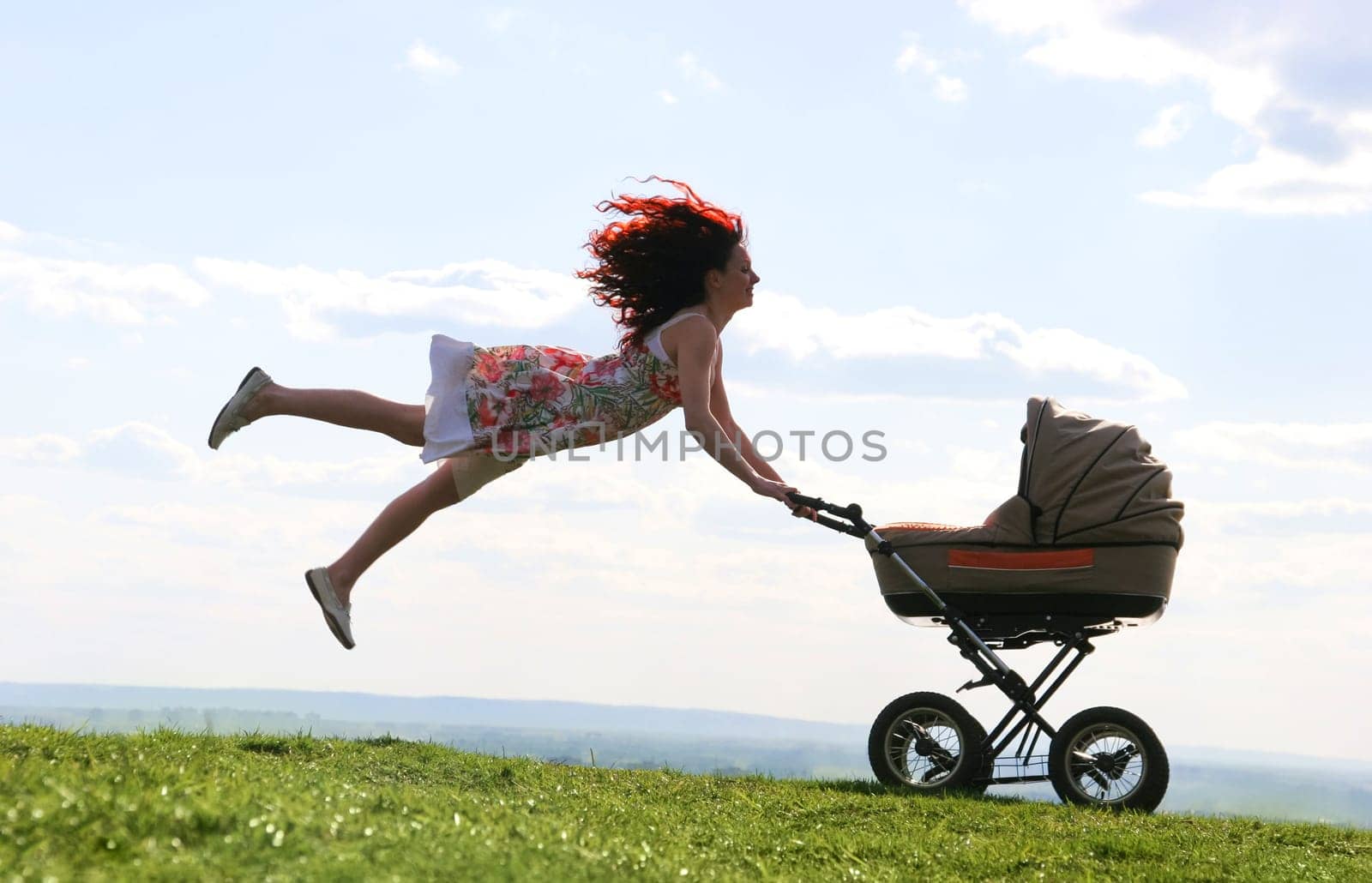 Joyful female jumping over green grassland while holding carriage with baby