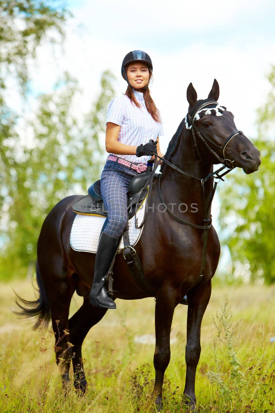 Image of happy female sitting on purebred horse and looking at camera outdoors
