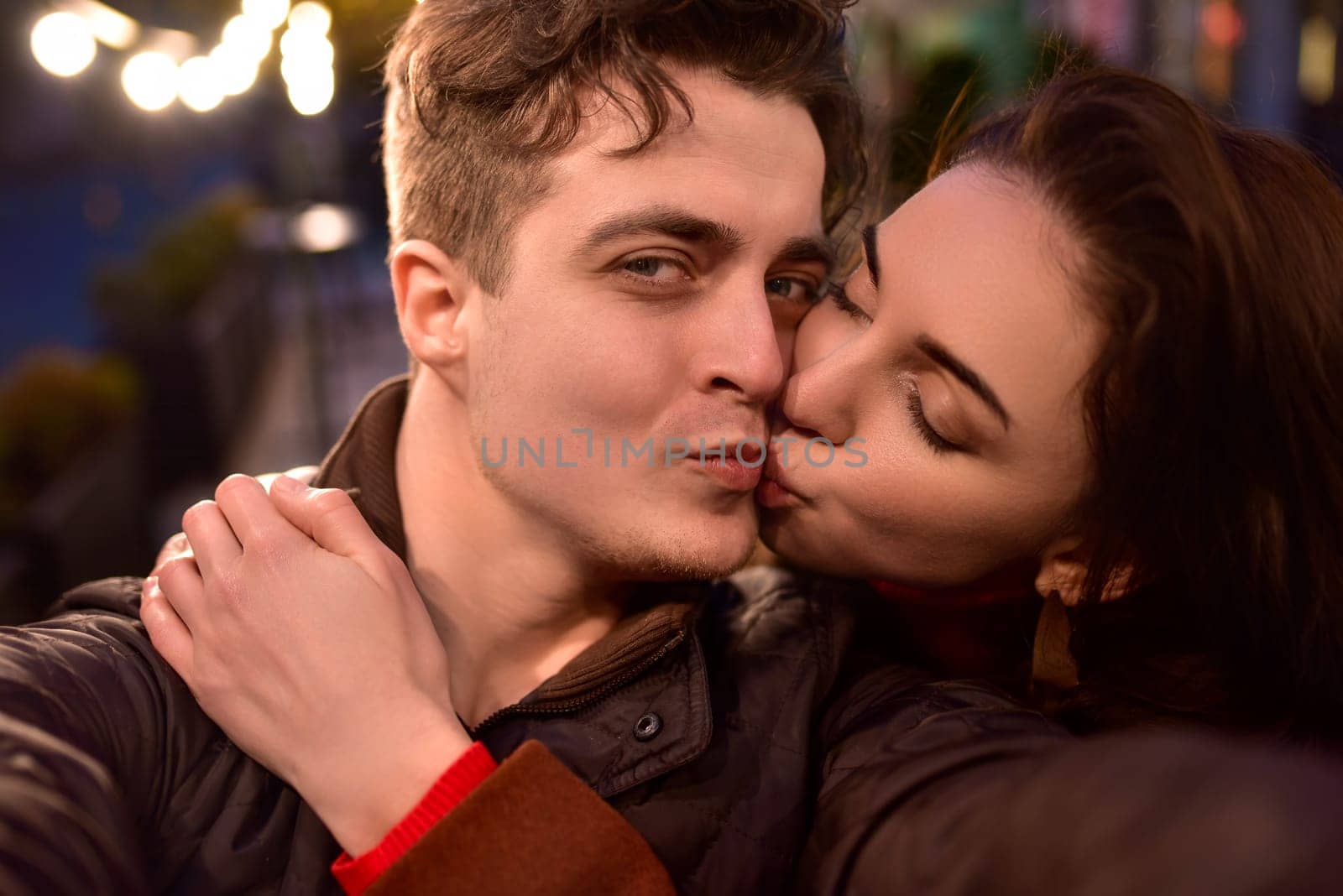 A young couple in love walks down a night street and takes selfies. The girl is kissing her boyfriend. by Nickstock