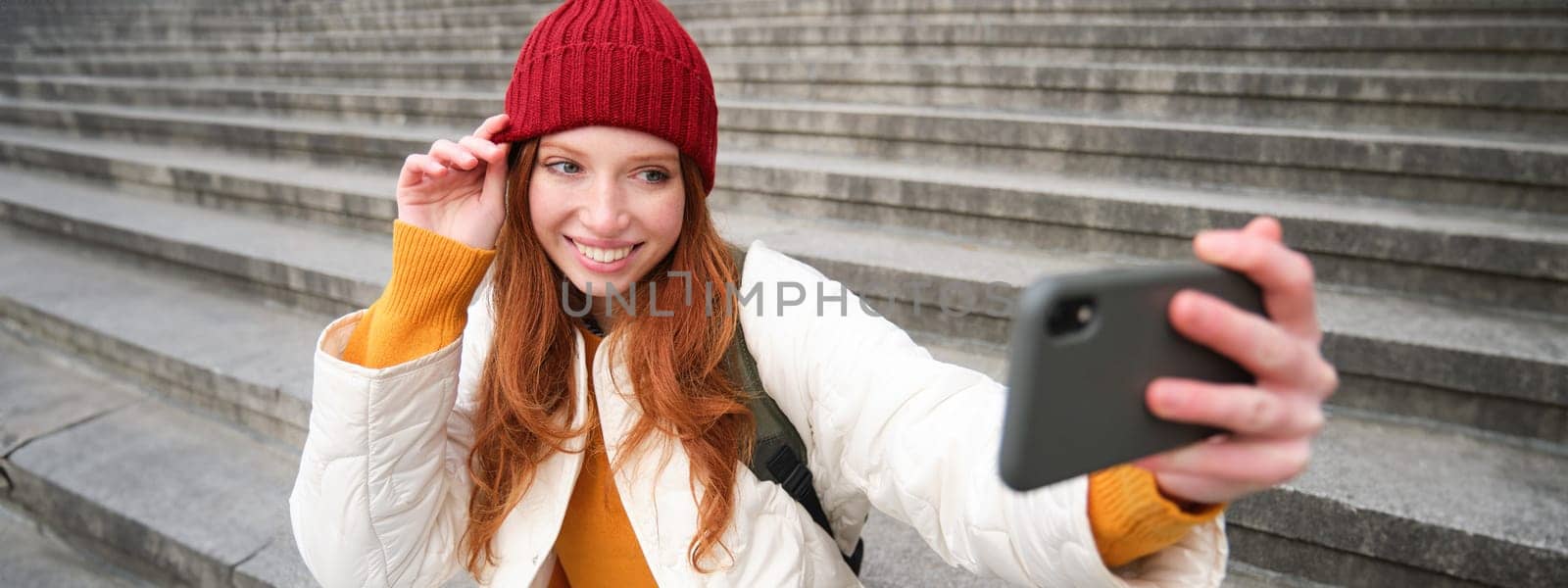 Stylish young girl in red hat, takes photos on smartphone camera, makes selfie as she sits on stairs near museum, posing for photo with app filter.