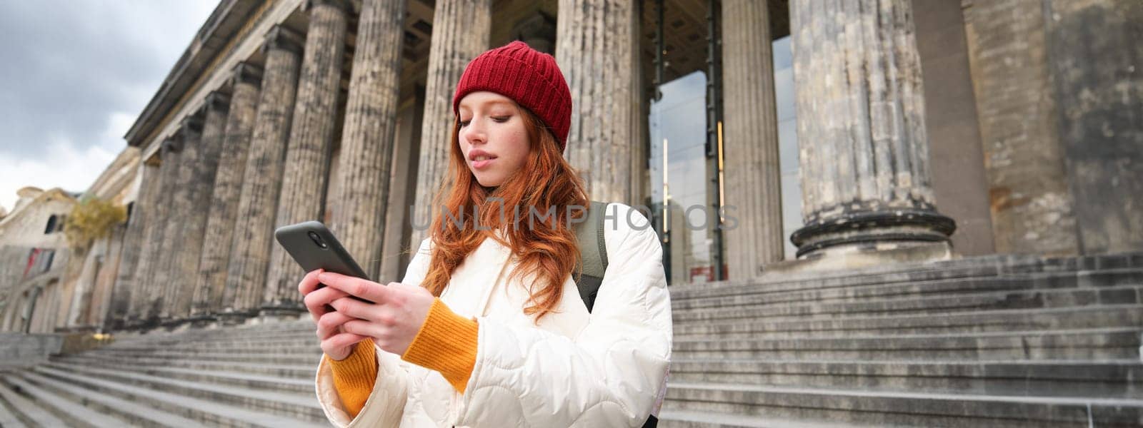 Portrait of young urban girl with backpack, using smartphone, walking around city, looking at map on mobile app, checking direction.